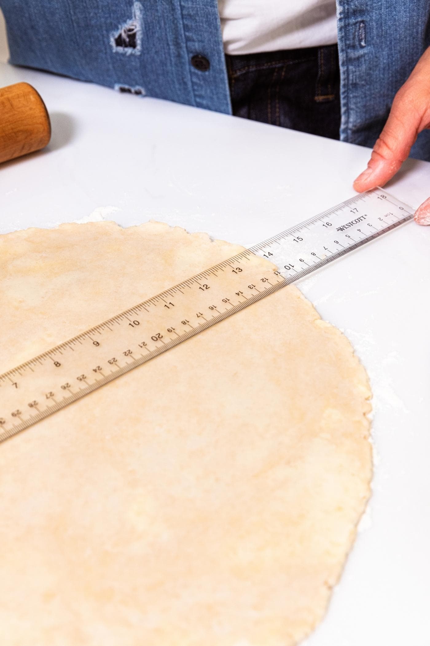 Hand placing ruler across dough that was rolled into a circle on white surface.