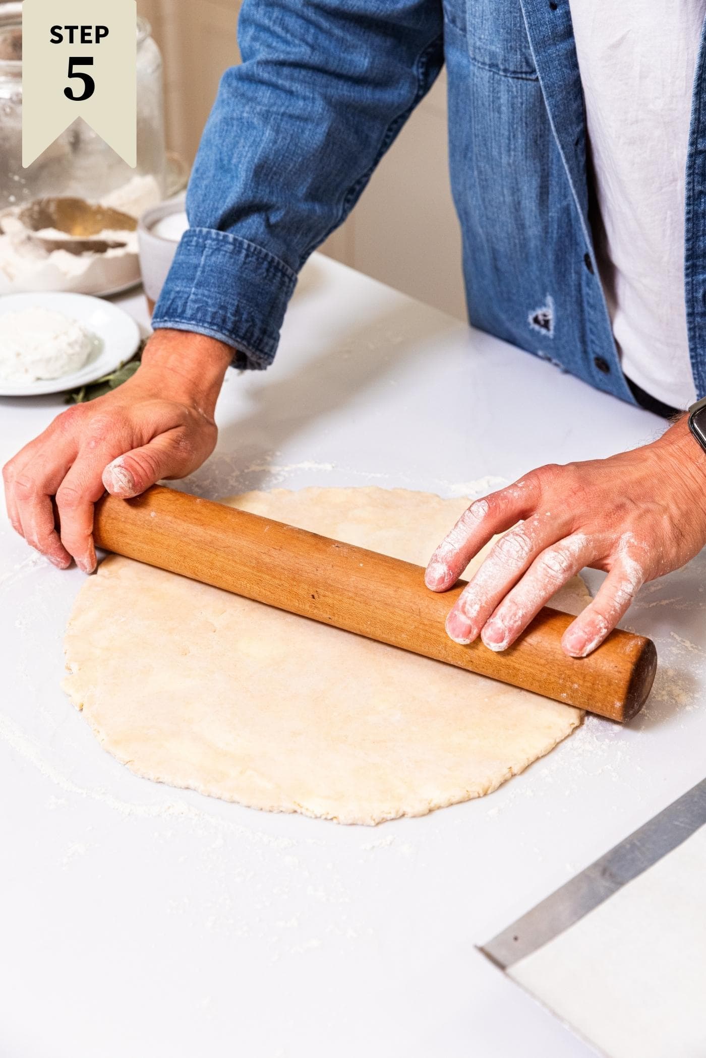 Two hands using wooden rolling pin to roll dough out into a circle on white surface.