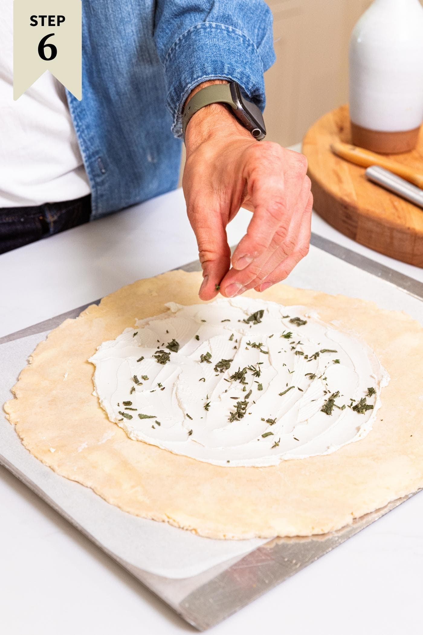 Hand sprinkling fresh herbs over the top of goat cheese spread over an unbaked piece of dough rolled out into a circle on a baking sheet.