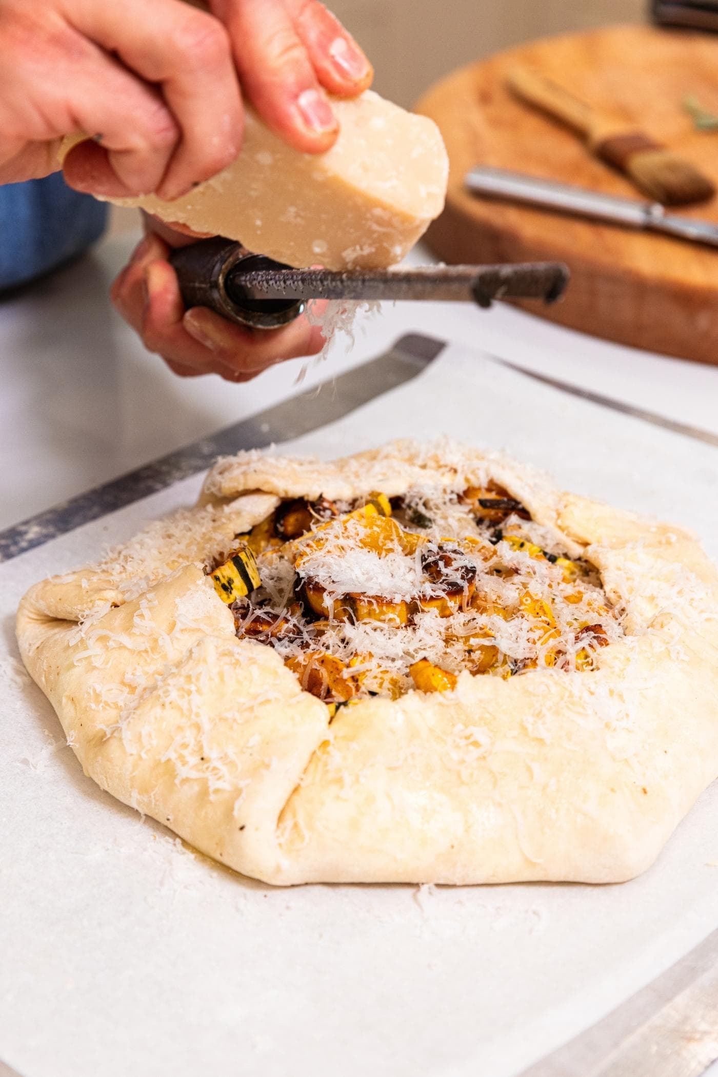 Hand using a grater to grate fresh Parmesan cheese on the top of an unbaked squash galette that has the sides folded up.