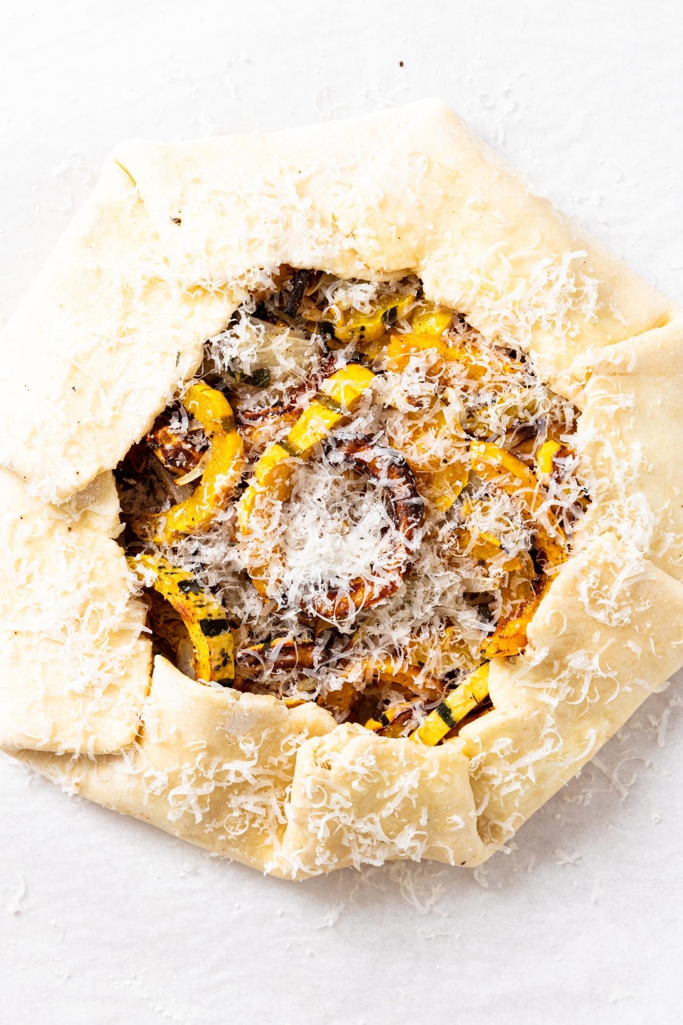 Unbaked roasted squash galette sitting on piece of parchment with grated Parmesan cheese all over the top.