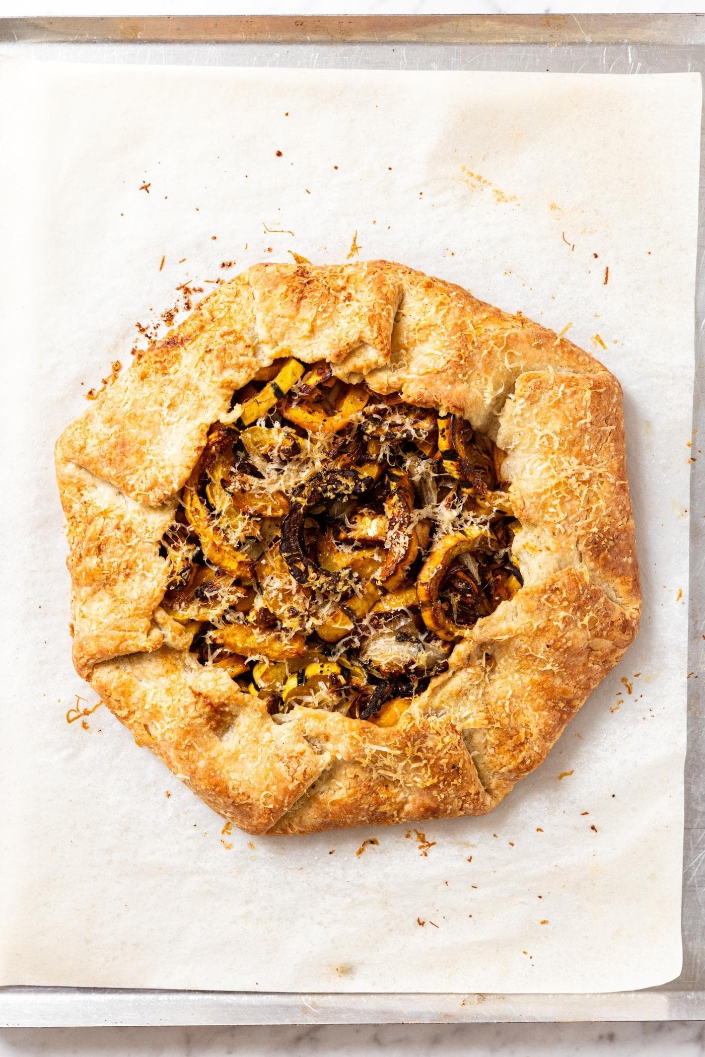 Baked roasted squash galette sitting on piece of parchment paper on baking sheet after coming out of the oven showing crispy golden brown texture.