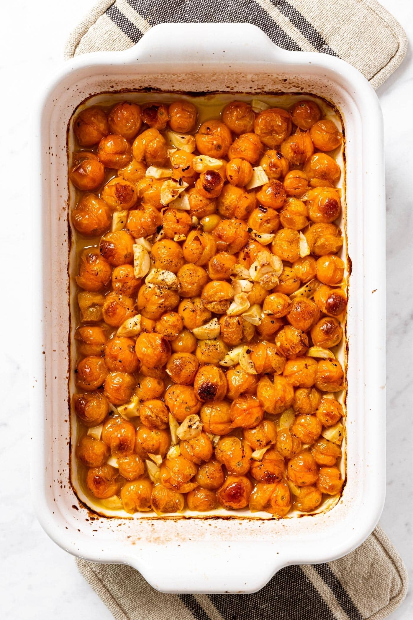 White baking dish filled with baked and burst Sungold tomatoes with garlic and shiny olive oil with baking mitts underneath.