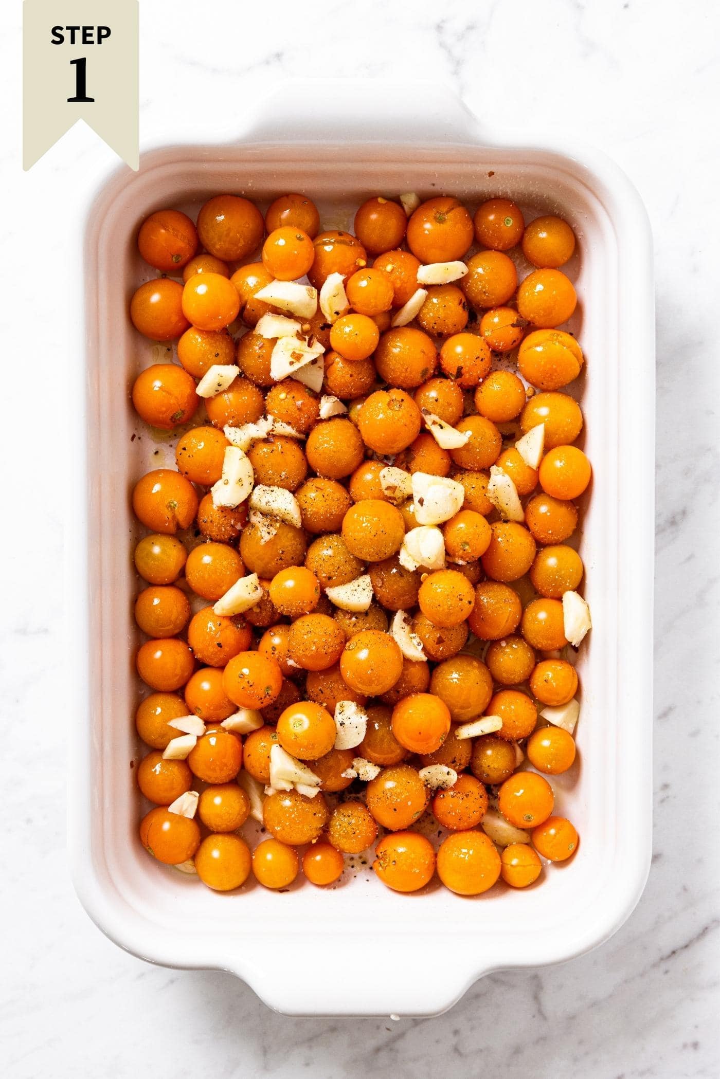 White baking dish with raw Sungold tomatoes sitting in the dish with garlic cloves and olive oil on top all on marble surface.