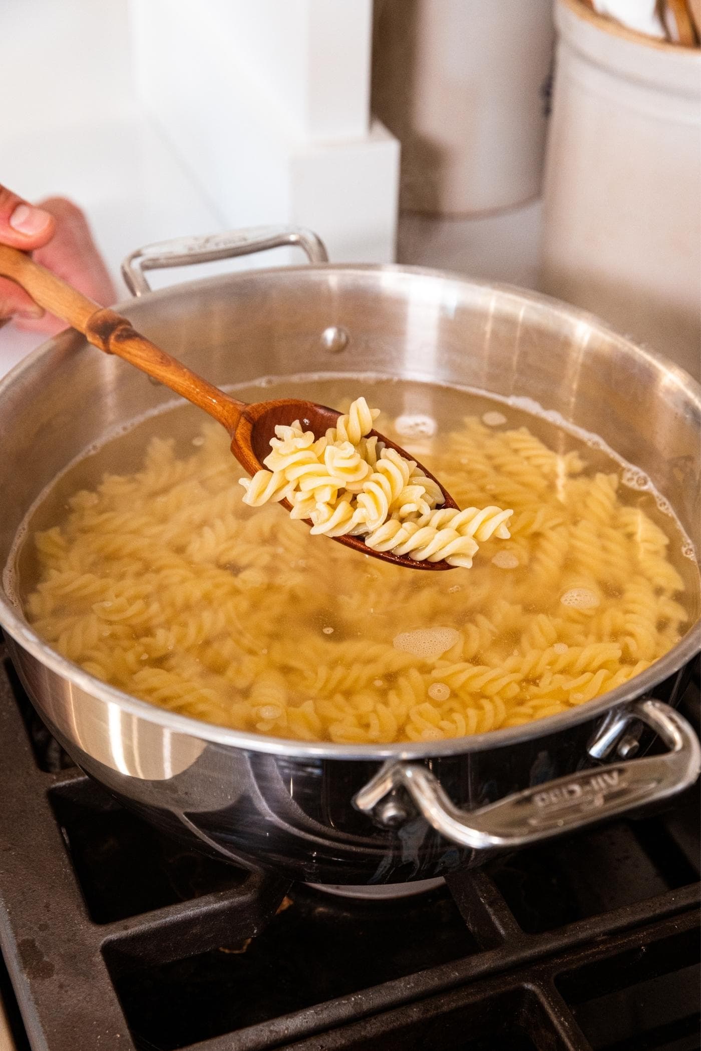 Hand holding wooden spoon above water cooking rotini pasta on stovetop.