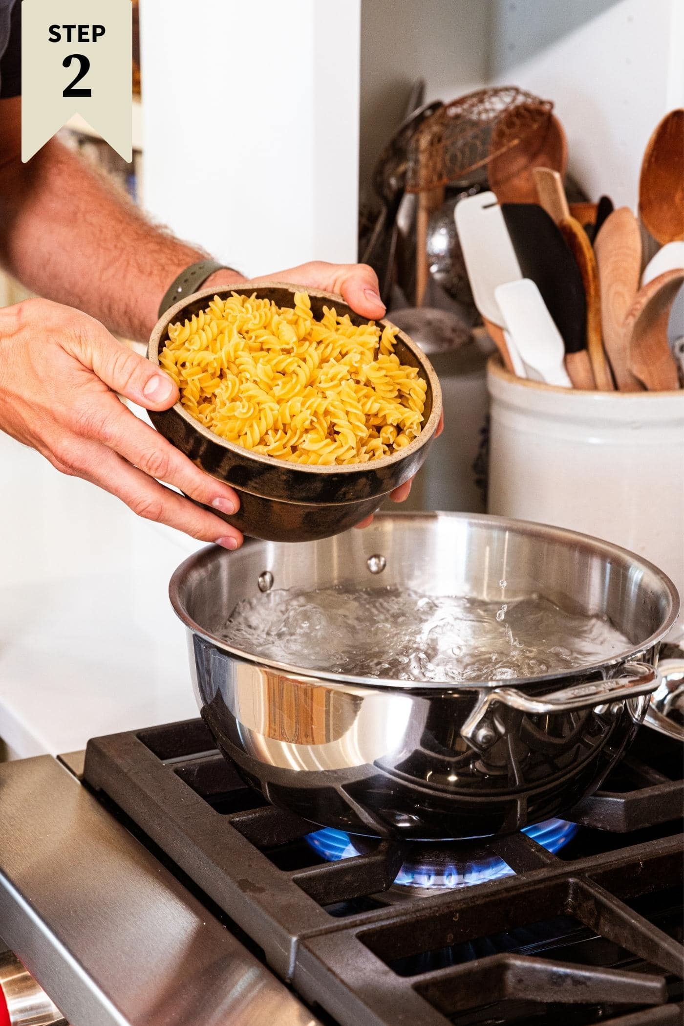 Two hands holding brown dish about to pour yellow rotini pasta into boiling water on stovetop.