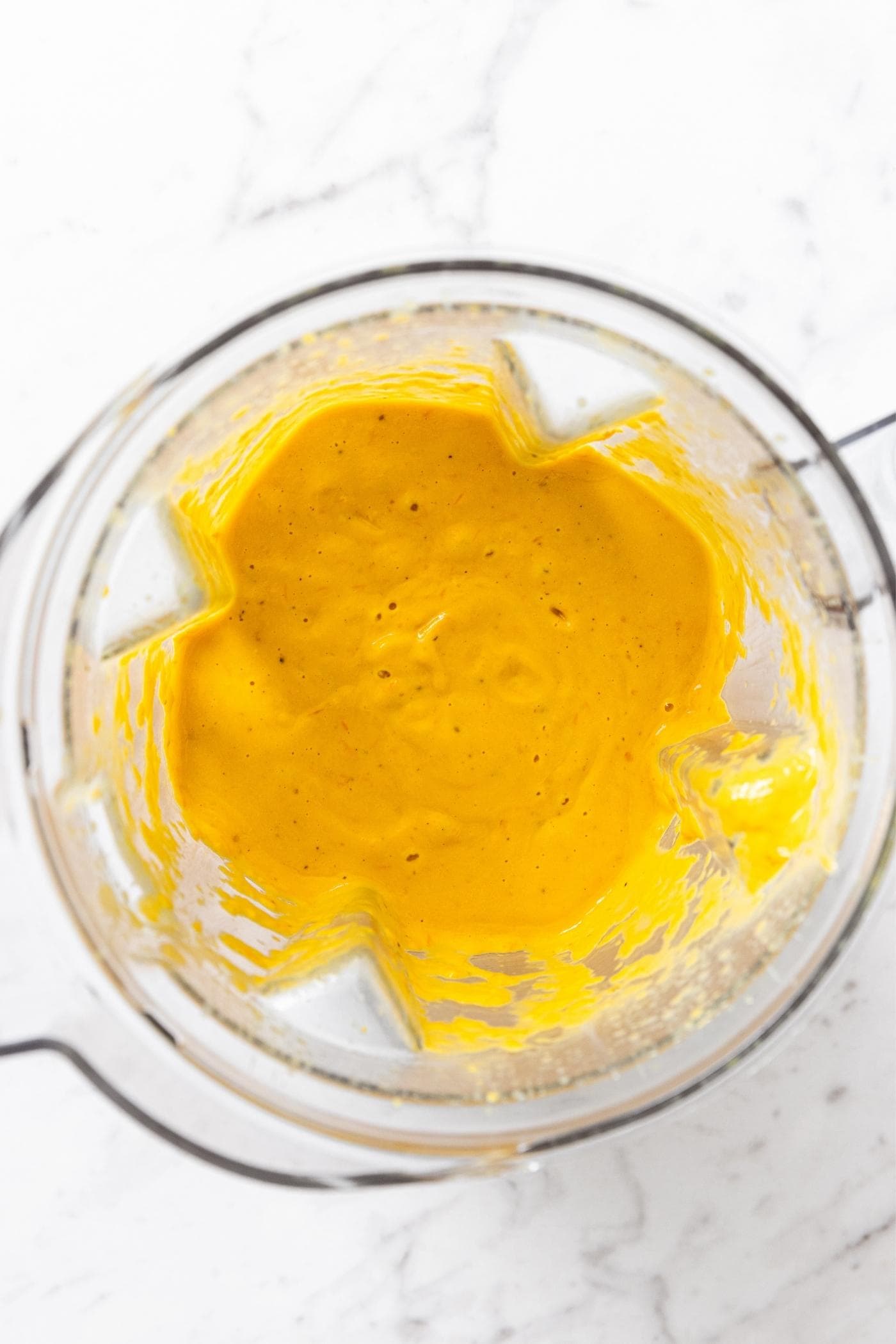 Blender container with blended yellow colored tomato sauce sitting on marble surface.