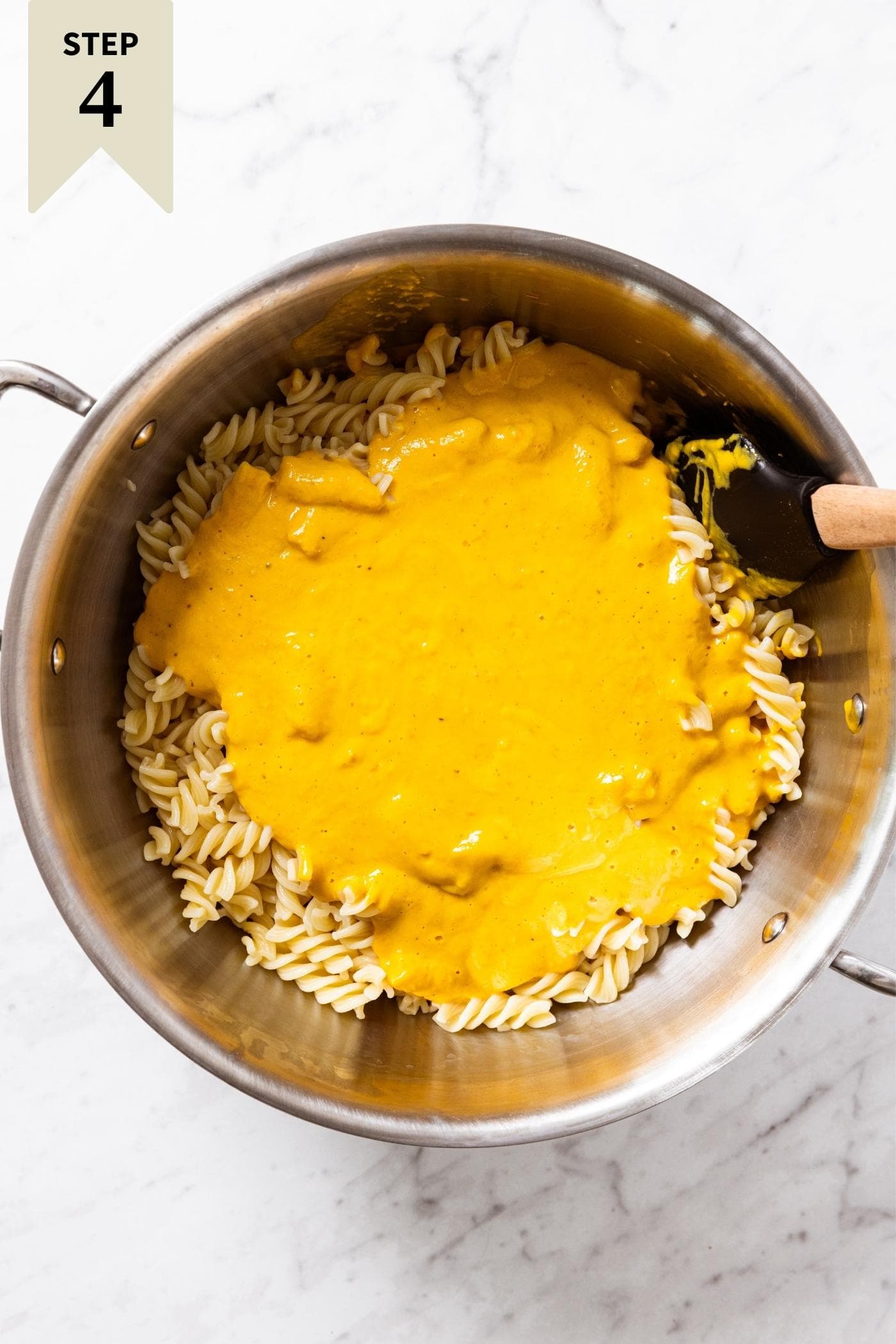 Spatula sitting in large metal pot filled with yellow pasta and yellow colored sauce before being mixed together all on marble surface.