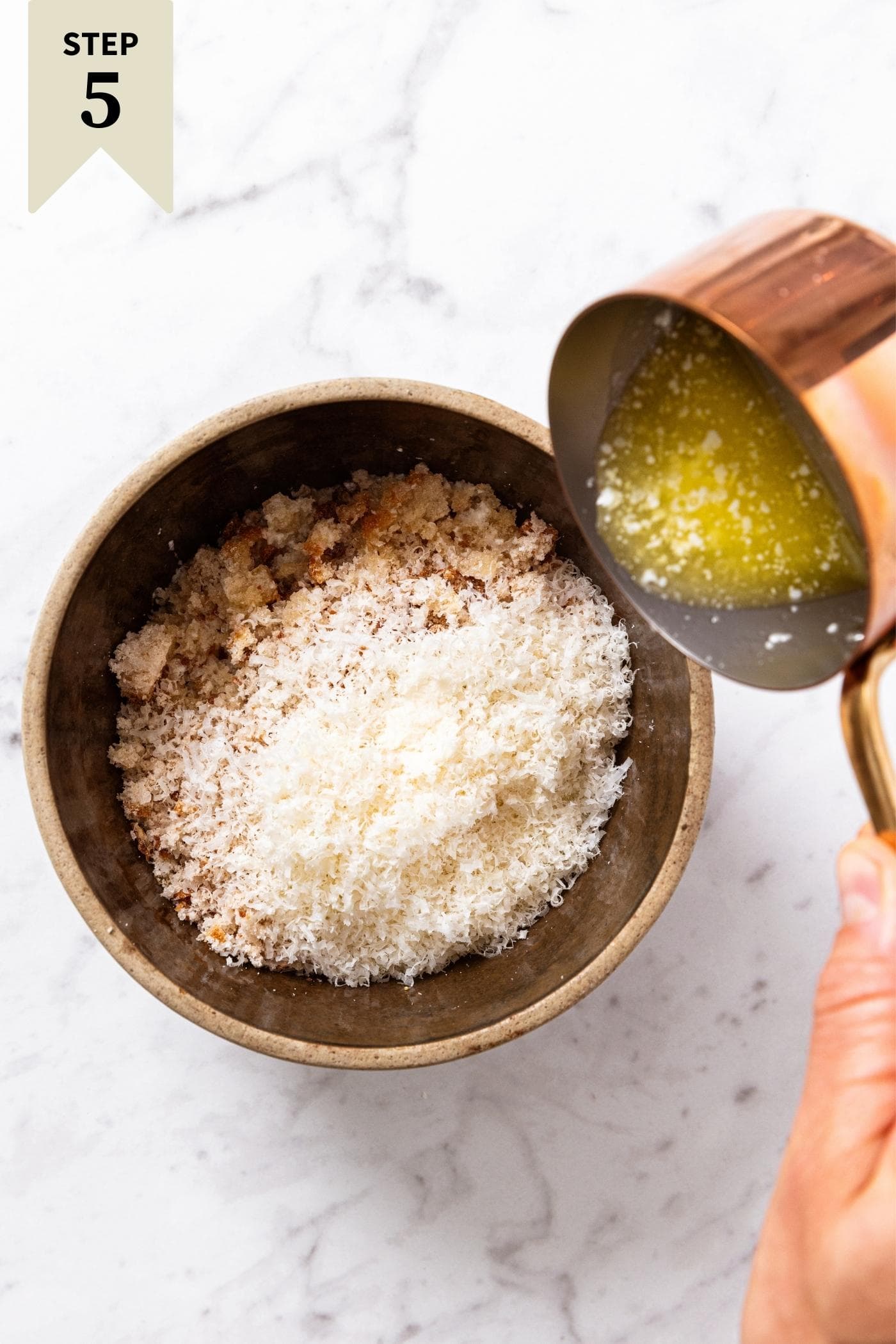 Hand pouring melted butter from small copper pot into a brown crock bowl filled with breadcrumbs and cheese.