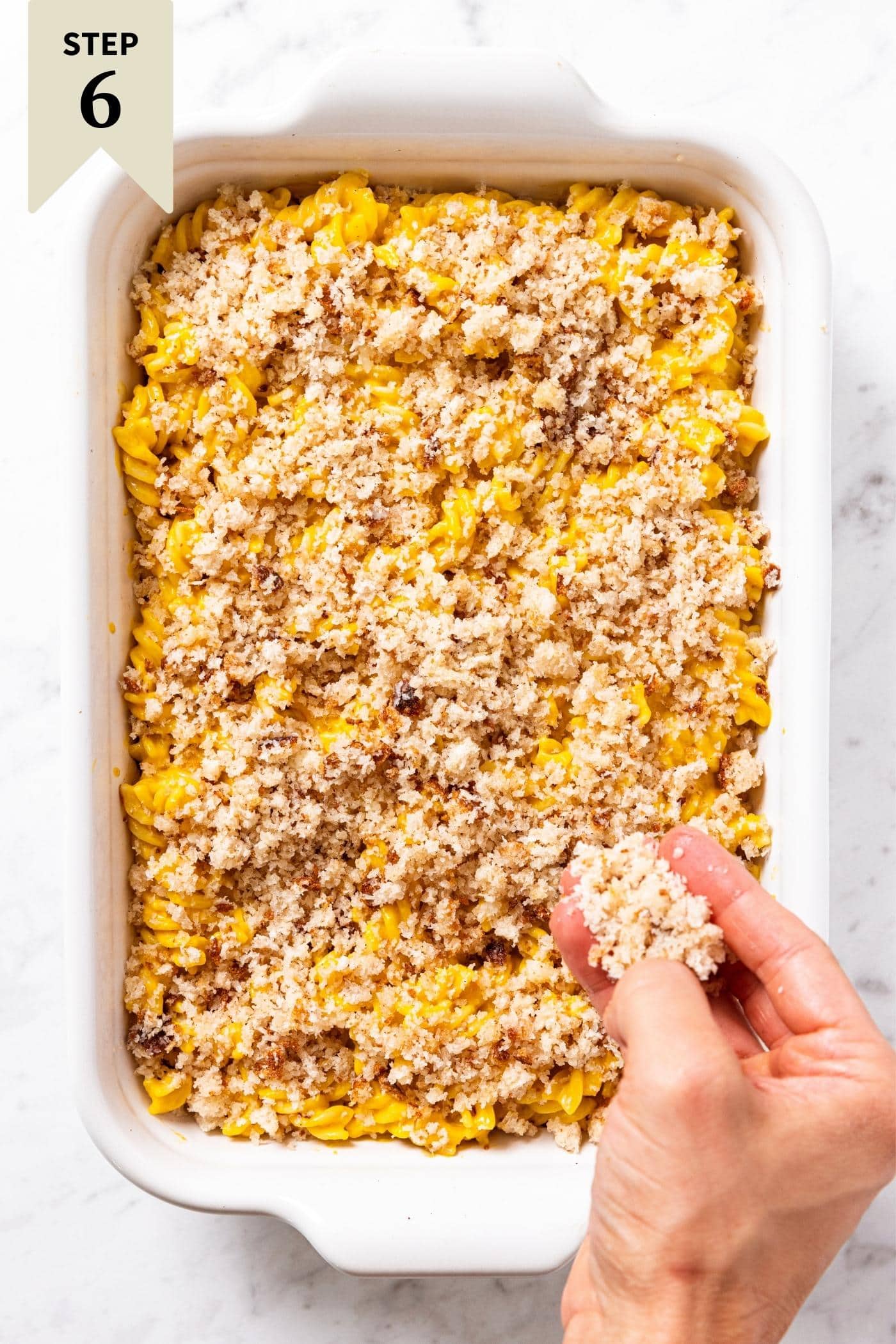 Hand sprinkling breadcrumb topping all over rotini pasta in white baking dish on marble surface. 