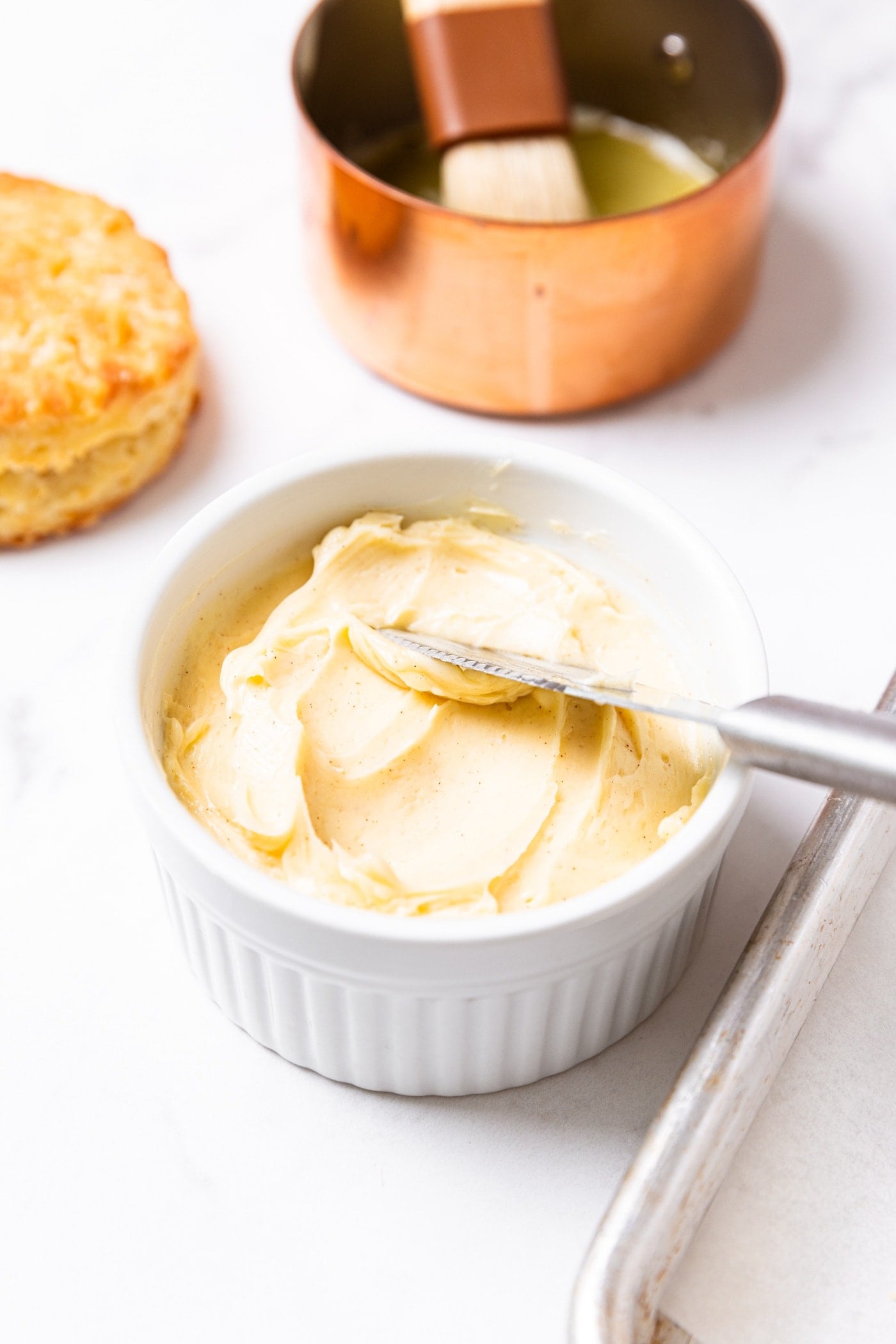 Butter knife sitting in small white bowl filled with cinnamon honey butter with copper pot in background as well as a golden baked biscuit.