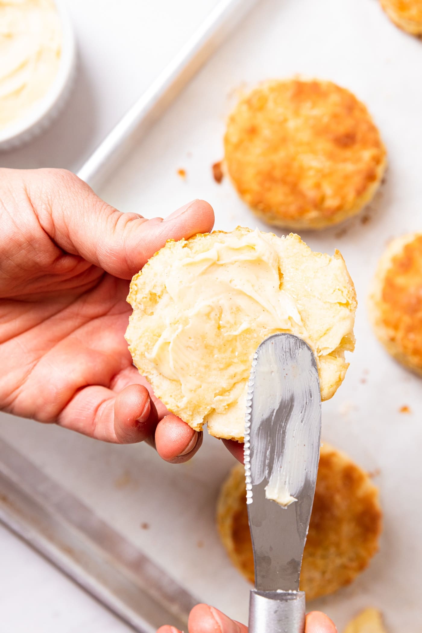 Hand using knife to spread honey butter on half of buttermilk biscuit with more biscuits below.
