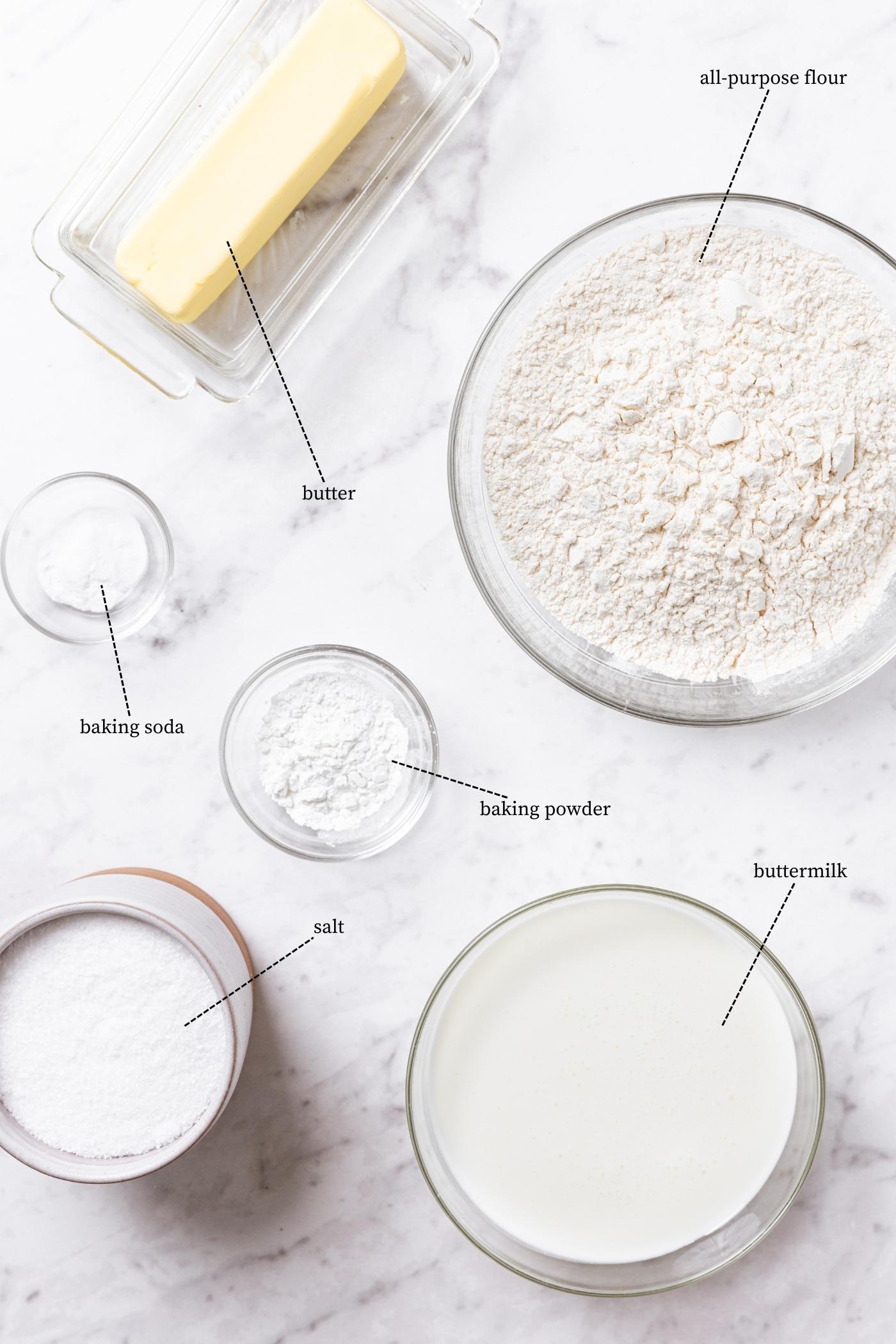 Marble surface with all ingredients needed to make buttermilk biscuits including flour, buttermilk, butter, baking powder and soda, and more.
