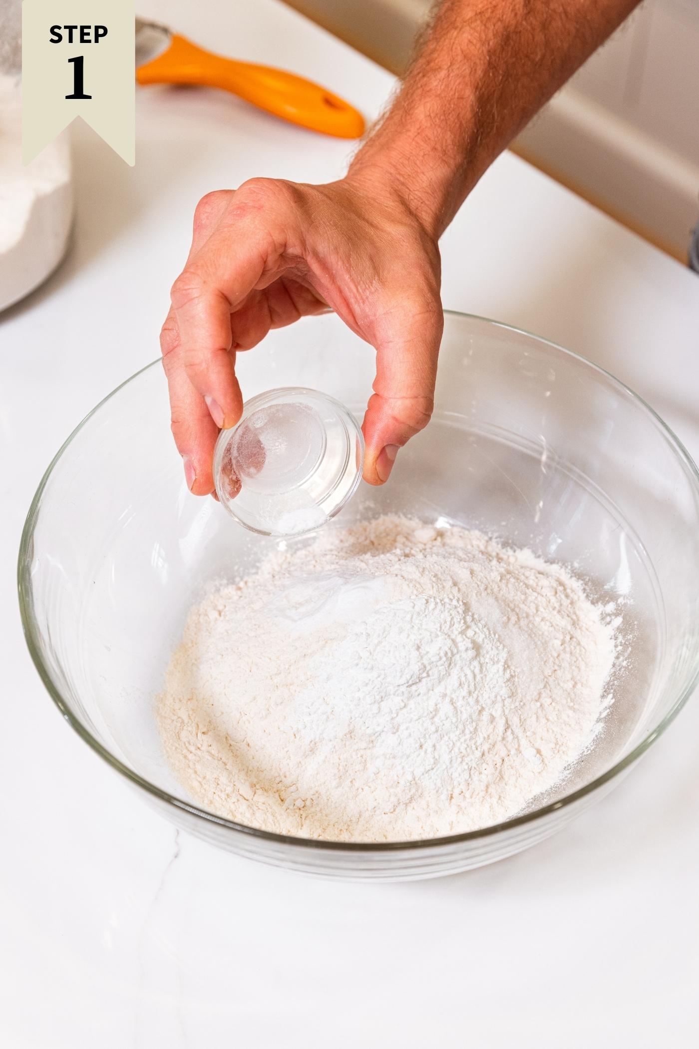 Hand pouring baking powder into glass bowl filled with flour and baking powder and salt all on white surface.