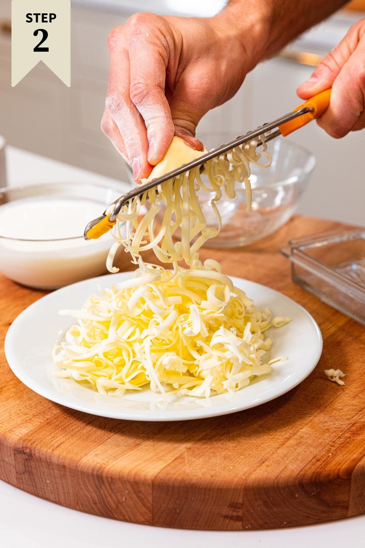 Hand using grater to grate butter into small pieces on white plate sitting on wood cutting board.