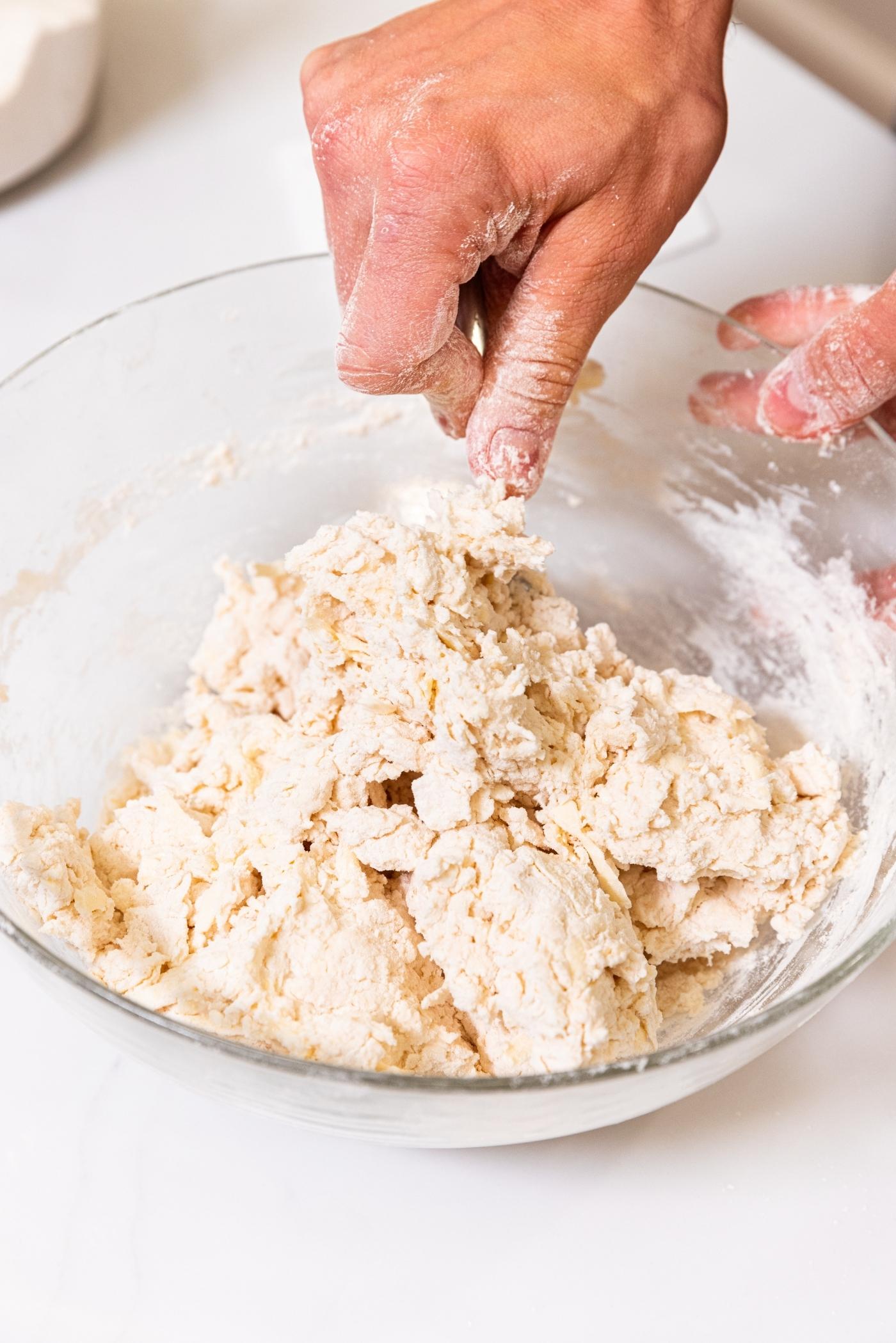 Hand using fork to mix together buttermilk with flour and butter mixture in glass bowl on white surface.