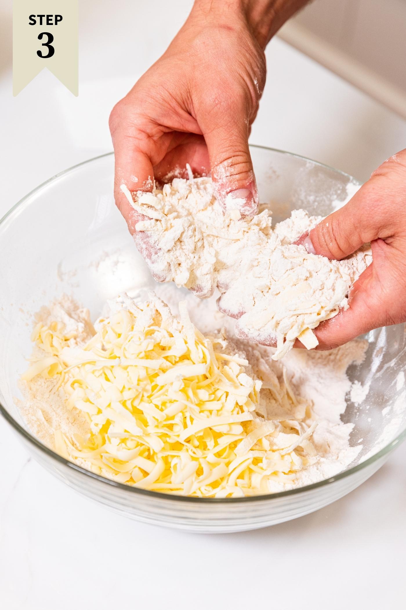 Two hands mixing together grated butter with flour mixture all in glass bowl on white surface.
