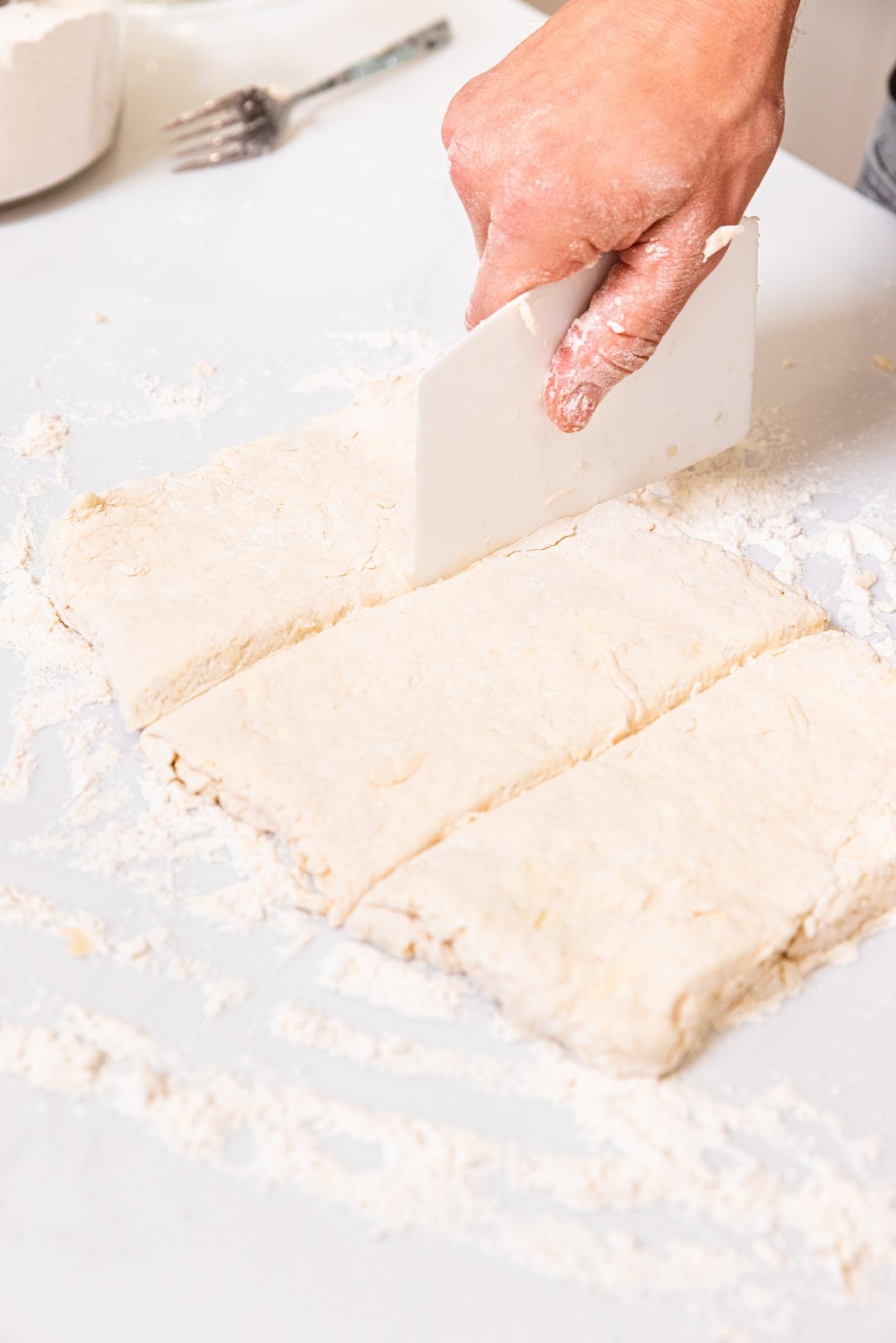 Hand using plastic piece to slice rolled out dough into thirds on floured white surface.