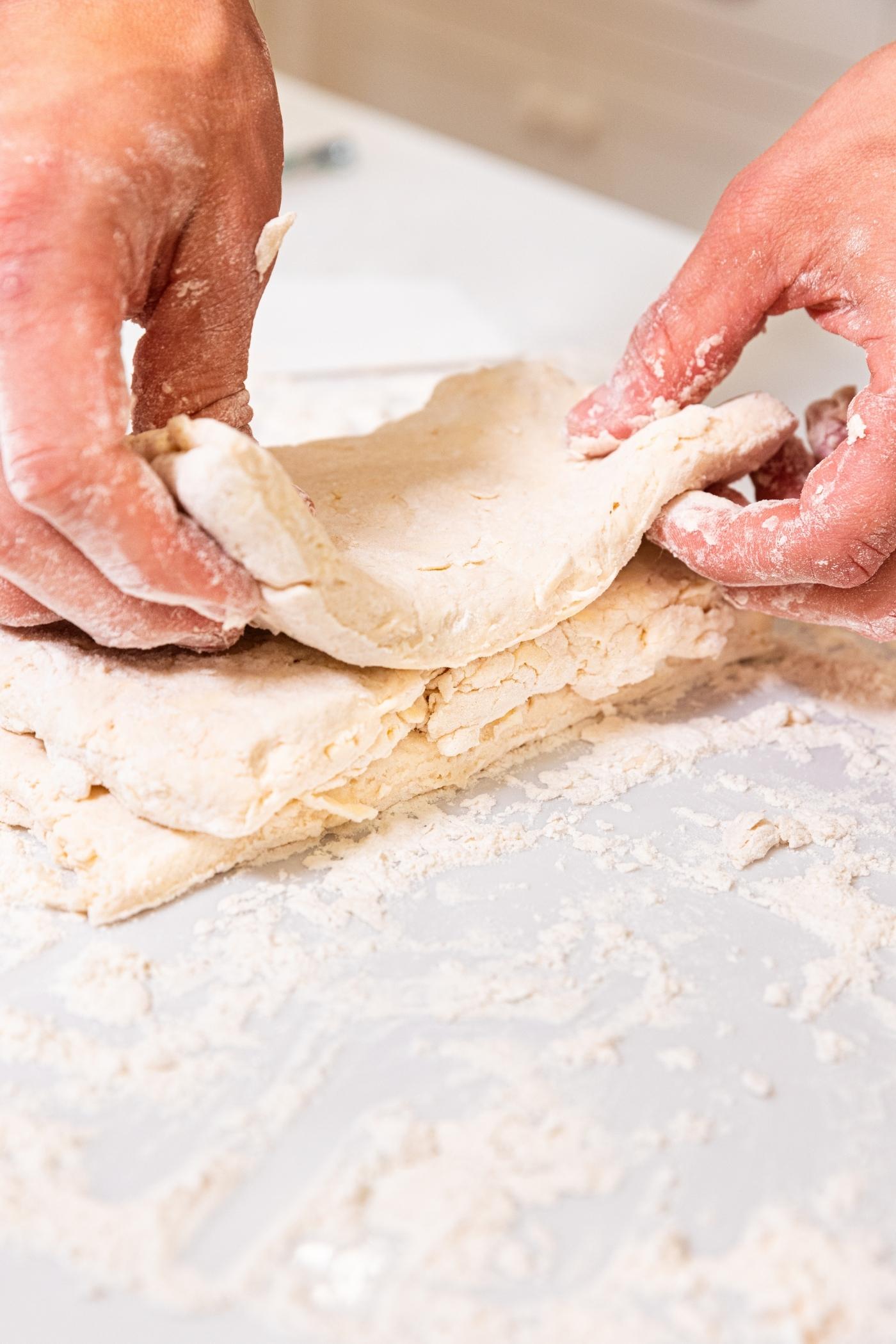 Hands placing pieces of dough on top of each other on floured white surface.