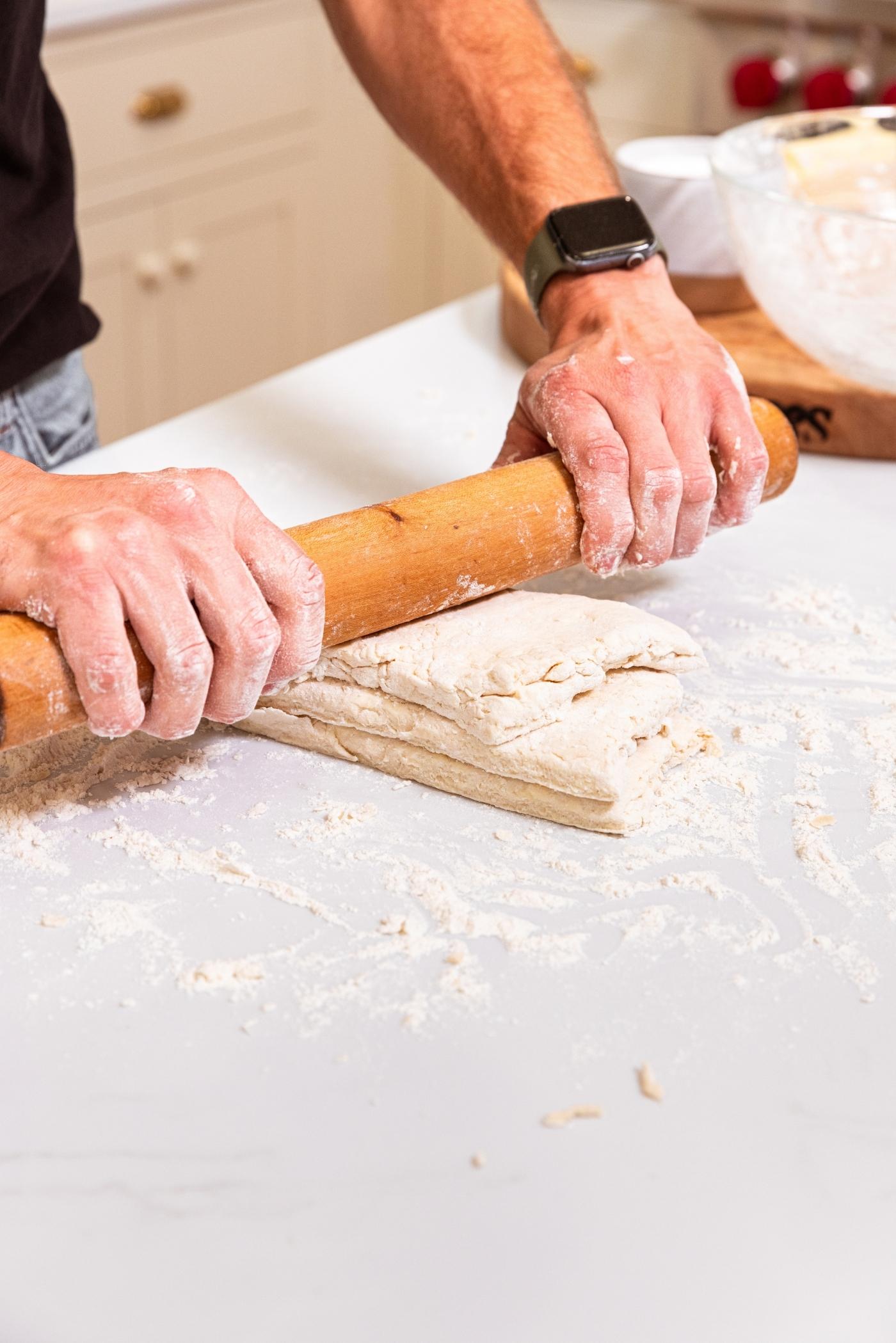 Two hands rolling out stacked pieces of dough on floured white surface.