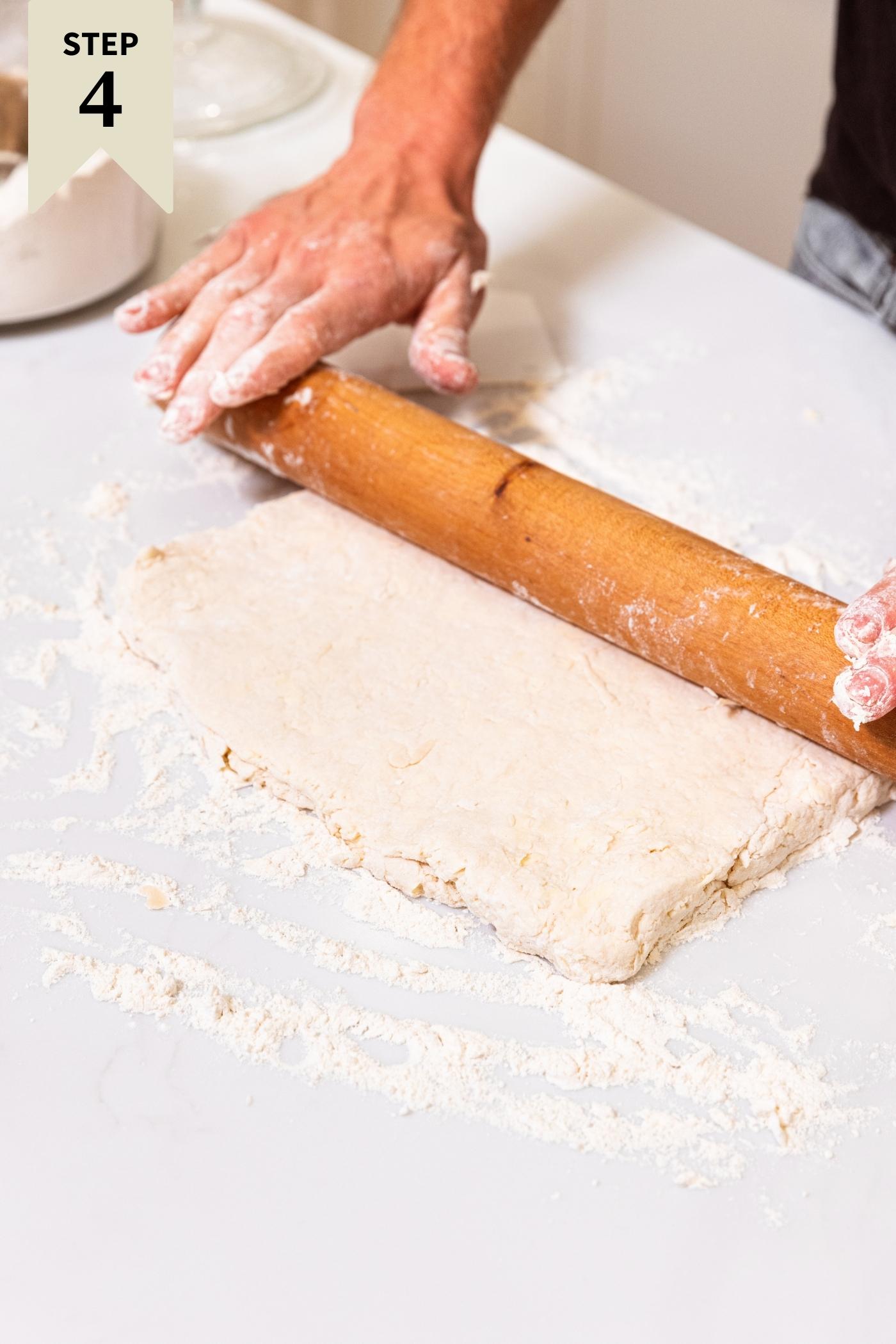 Hand on wooden rolling pin rolling out rectangular dough on floured white surface.