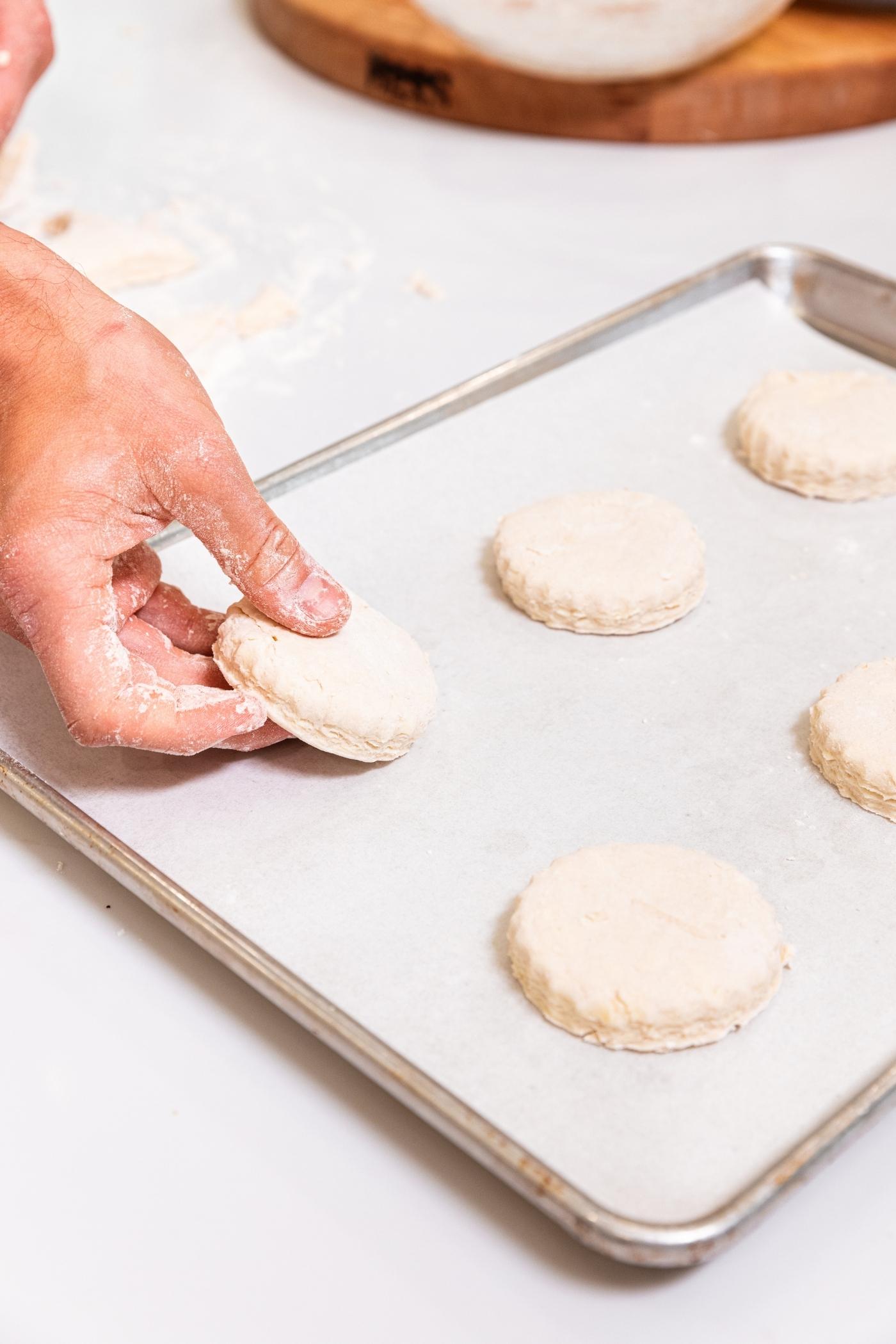 Hand placing a cut out biscuit onto a parchment-lined baking sheet on white surface with more biscuits all around.