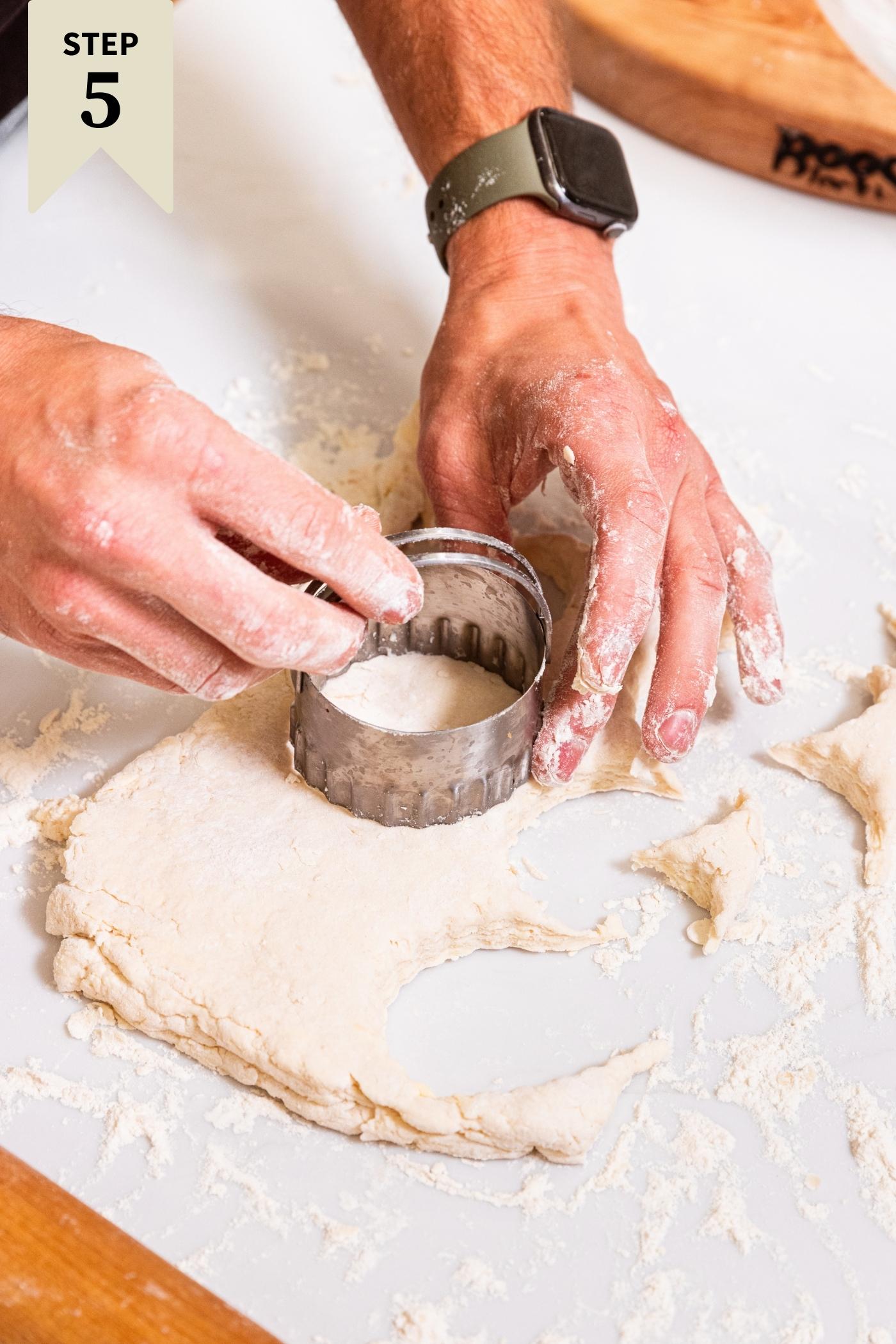 Hand cutting out biscuit with a biscuit butter from rectangle of dough on floured white surface.