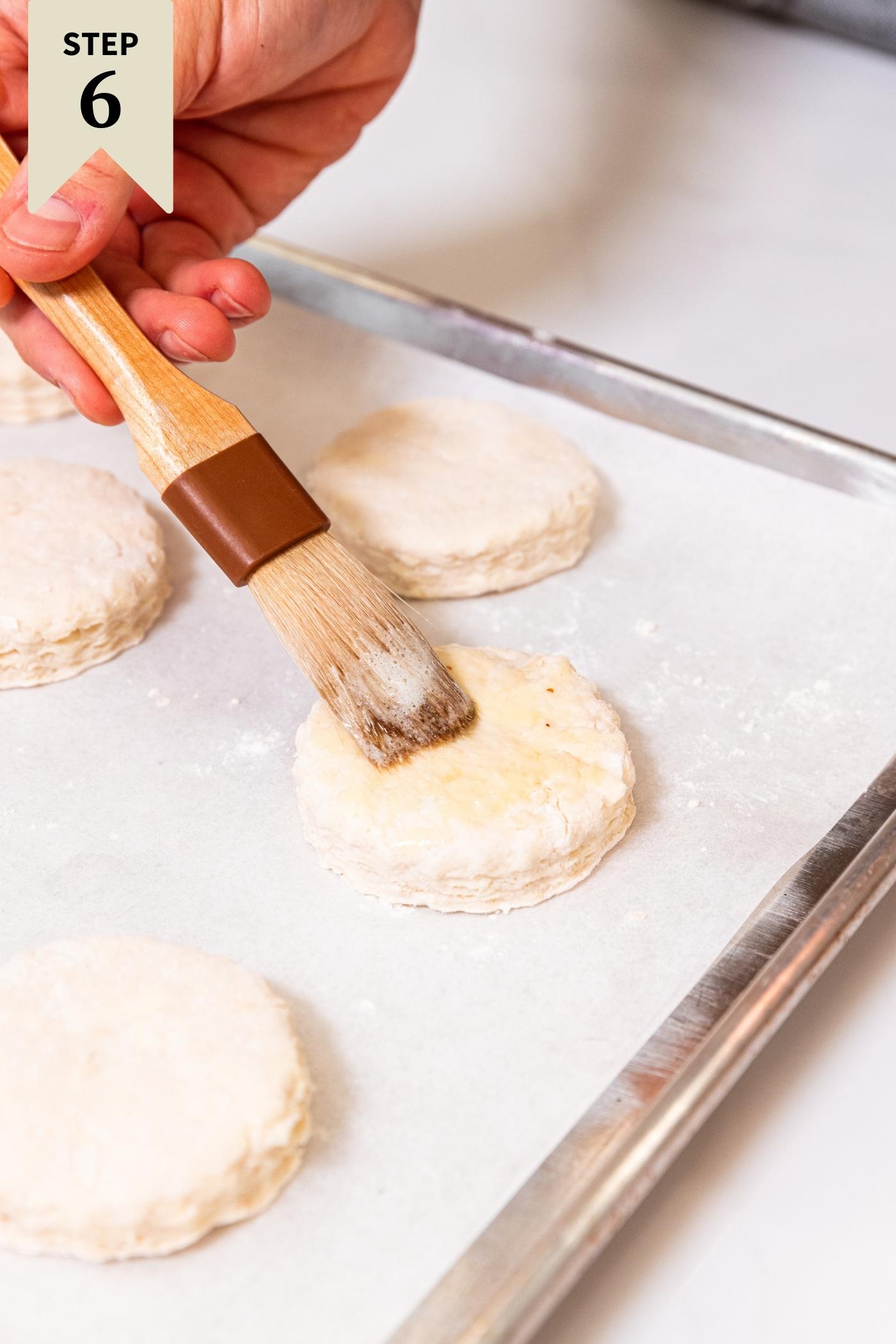 Hand brushing melted butter onto the top of an uncooked biscuits on baking sheet with brush.