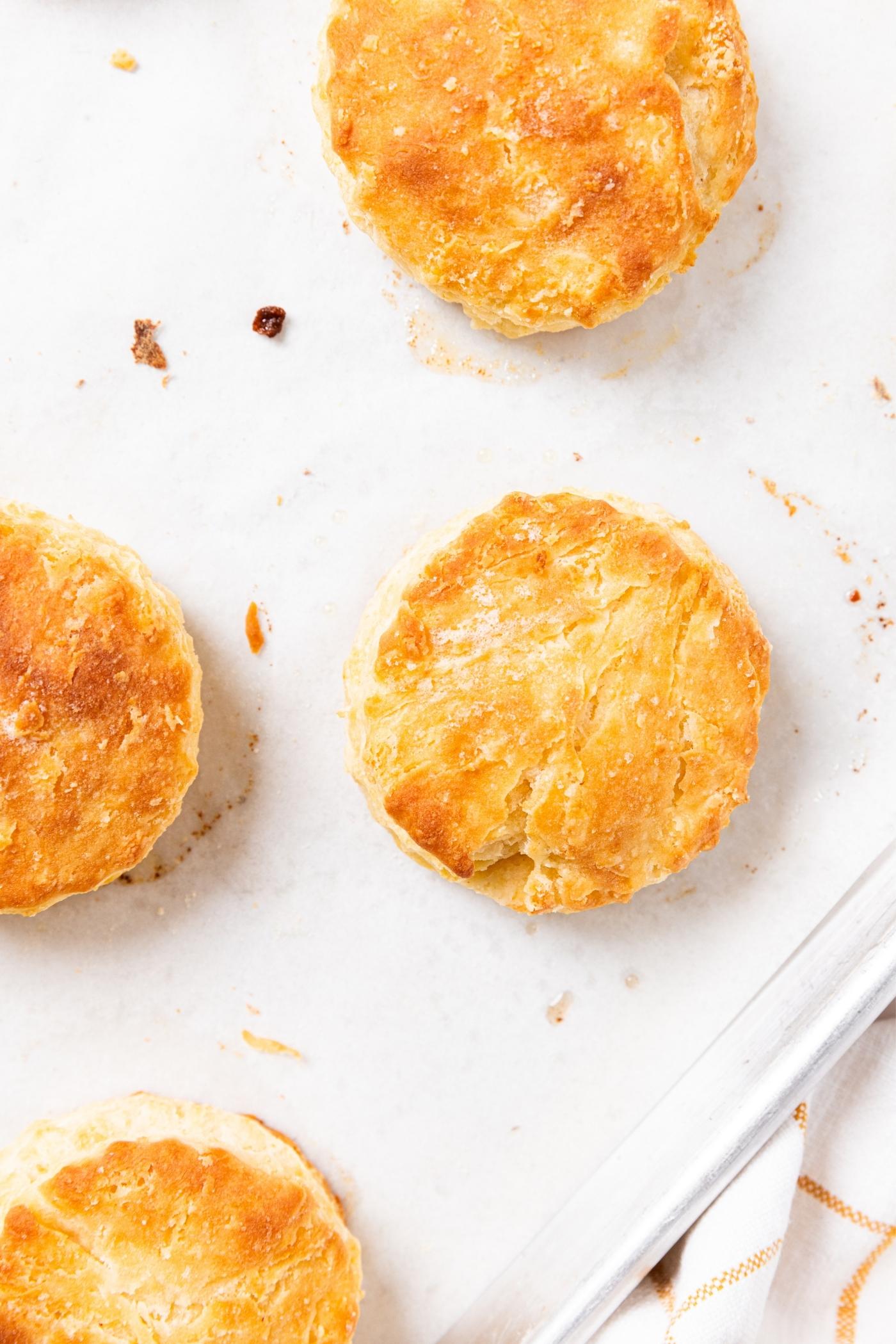 Baked buttermilk biscuits sitting on piece of white parchment on baking sheet after coming out of the oven.