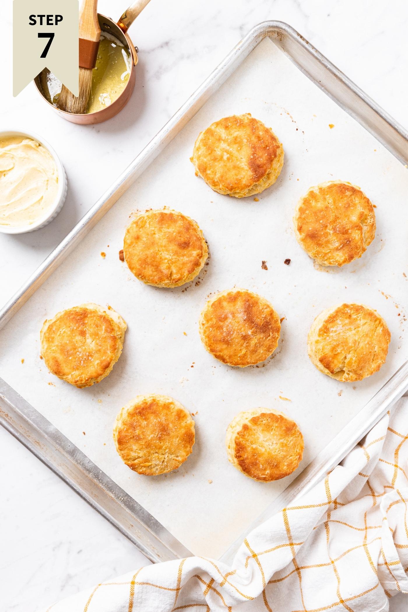 Baked biscuits sitting on parchment-lined baking sheet on marble surface with honey butter in white container and melted butter in copper pot.