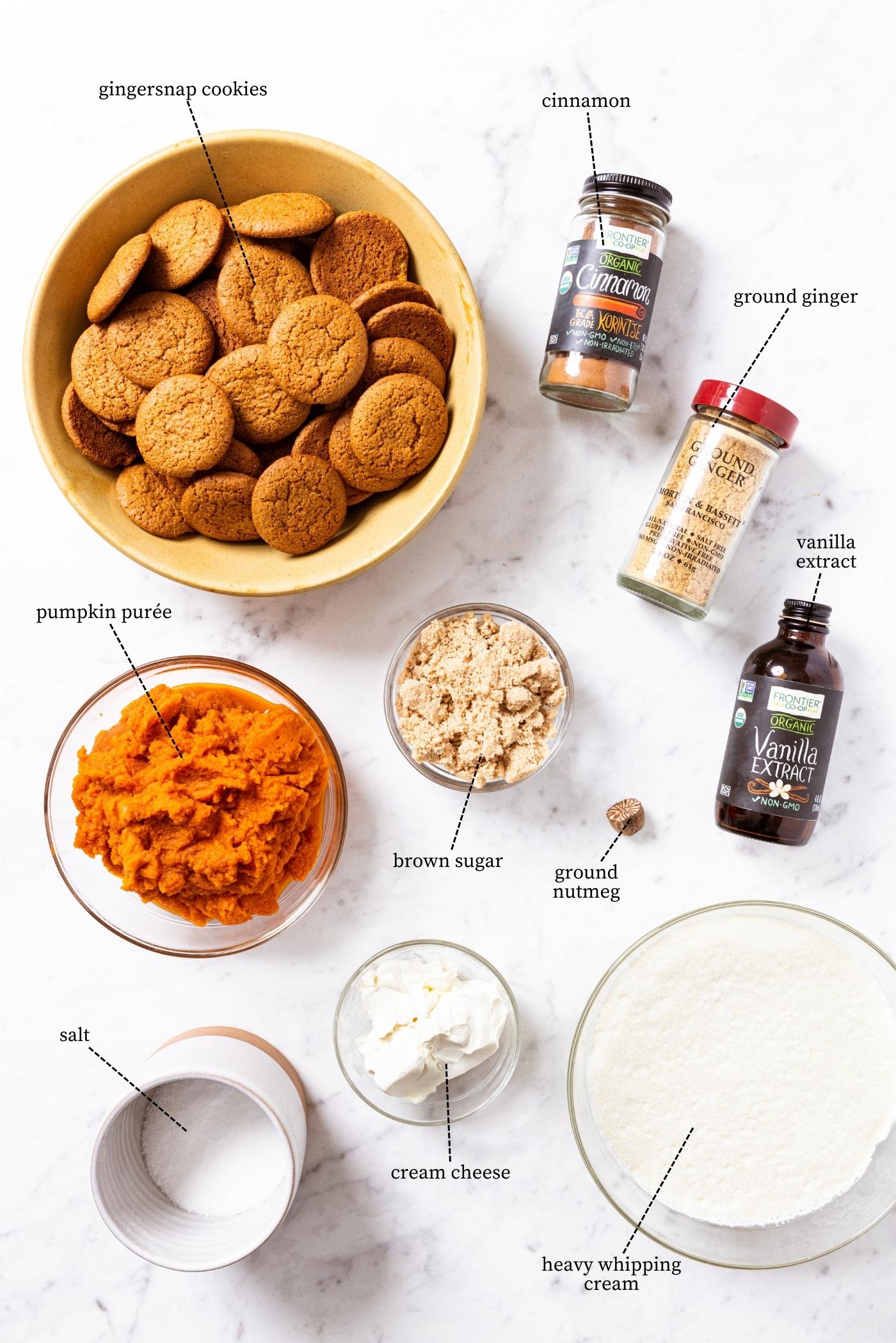 White marble surface filled with all ingredients needed to make pumpkin icebox cake including pumpkin puree, gingersnap cookies, spices, brown sugar, cream, and more.