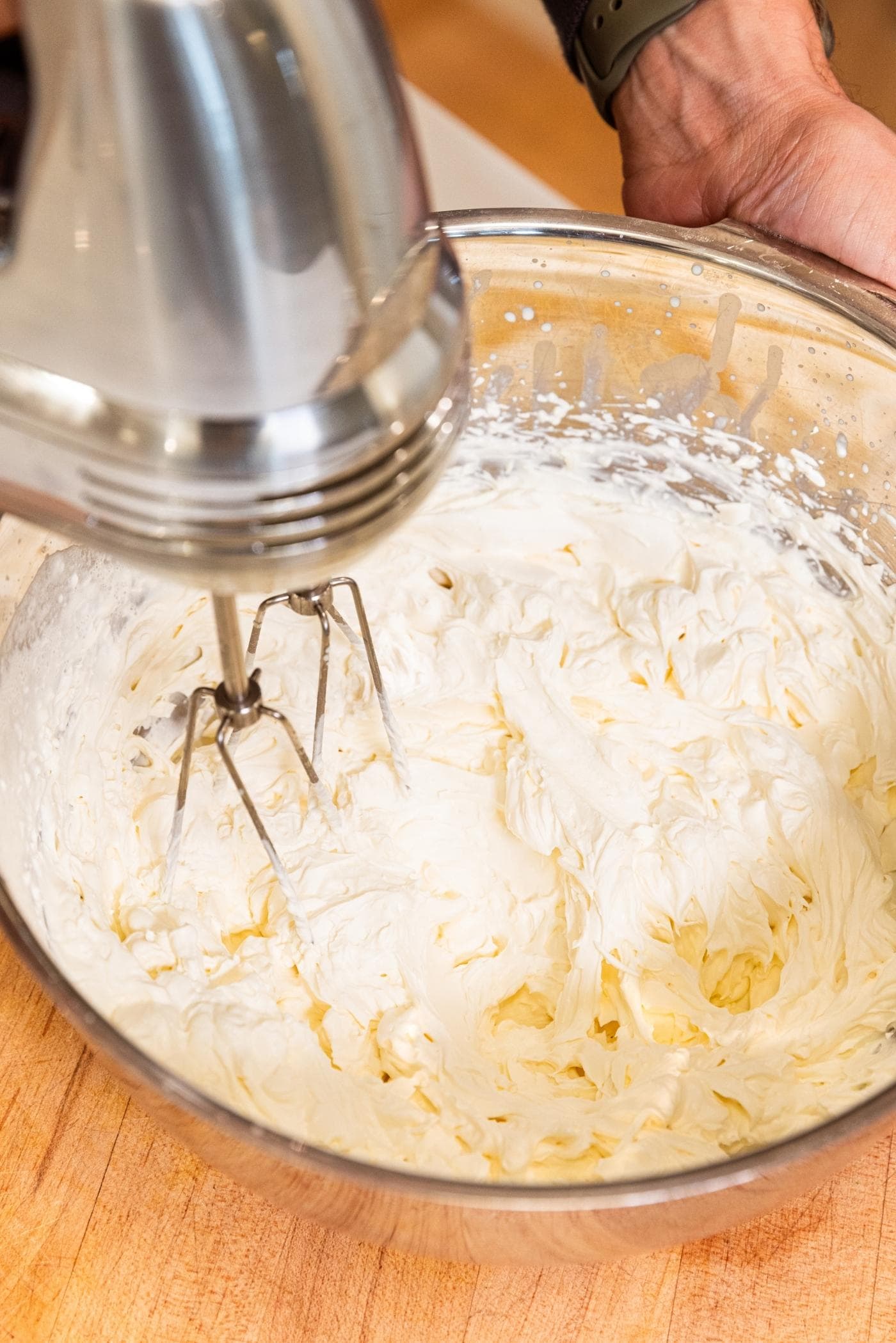 Hand holding metal bowl filled with whipped cream after being whipped by hand mixer with metal cutting board underneath.