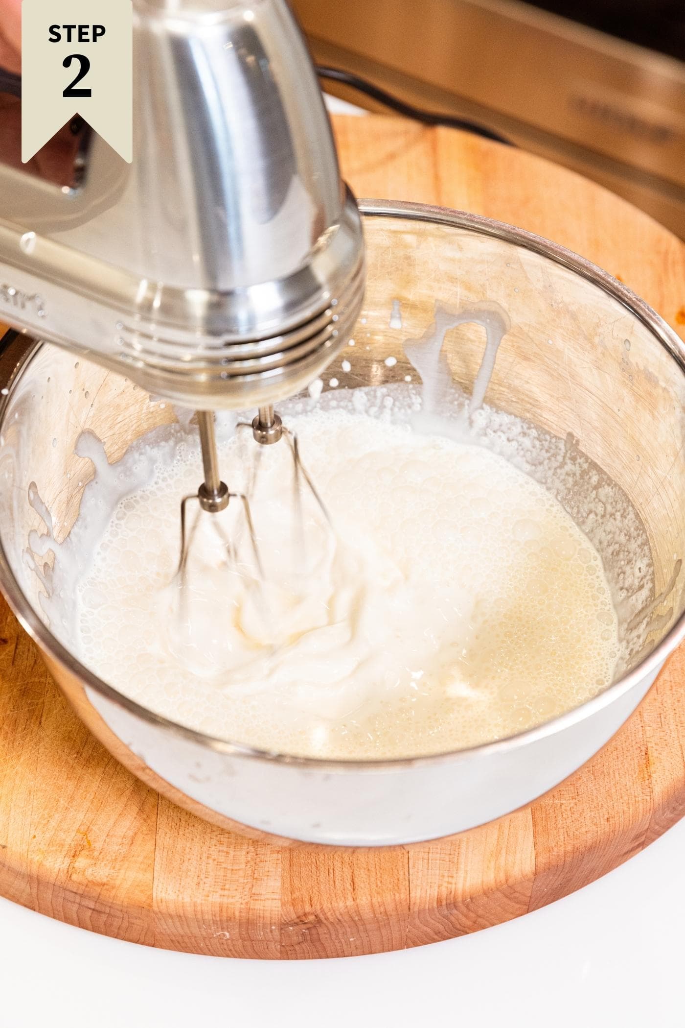 Metal bowl sitting on wood cutting board filled with heavy cream being whipped into whipped cream with a metal hand mixer.
