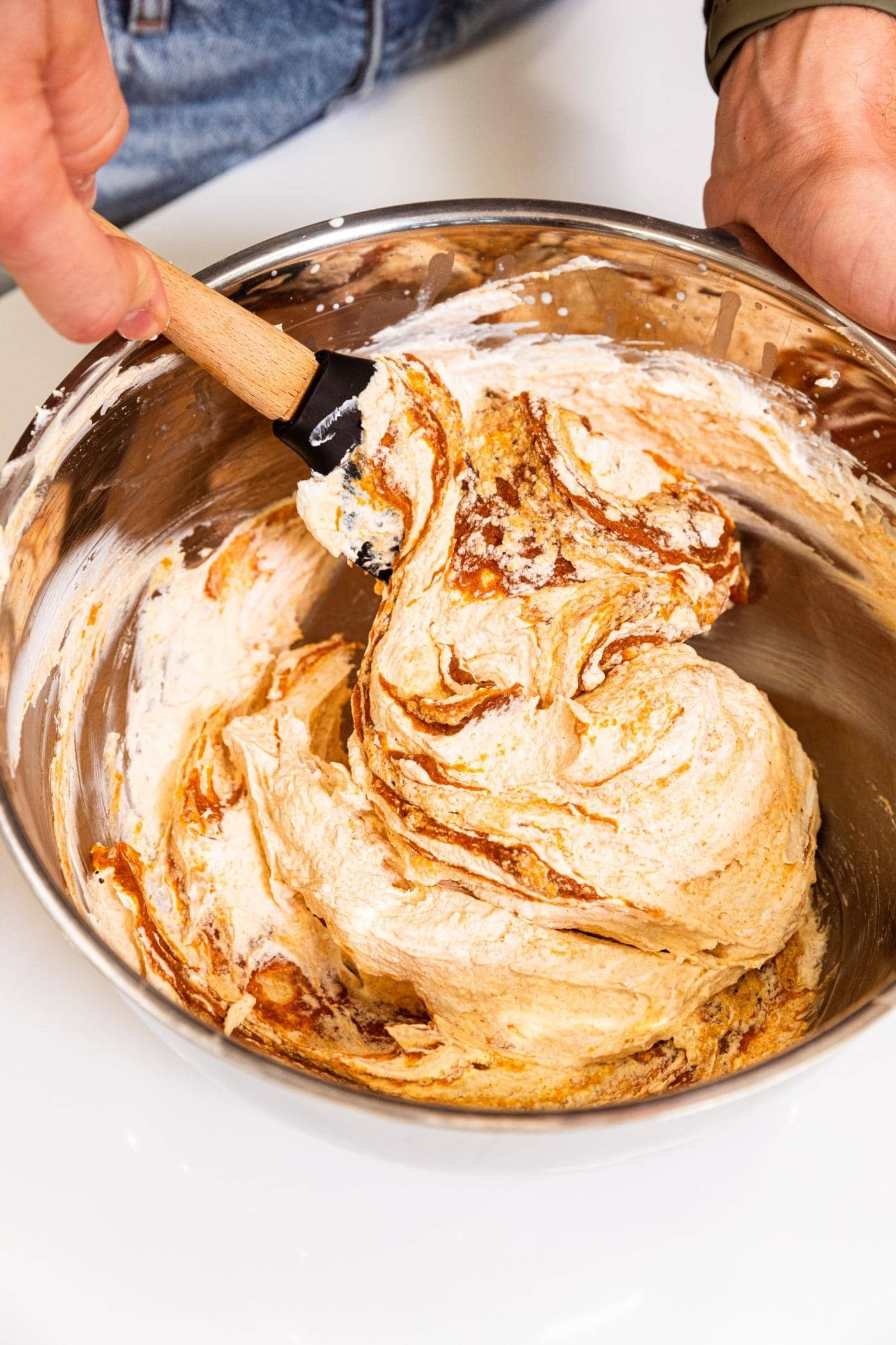 Plastic spatula mixing together pumpkin mixture with whipped cream in metal bowl with white surface underneath.