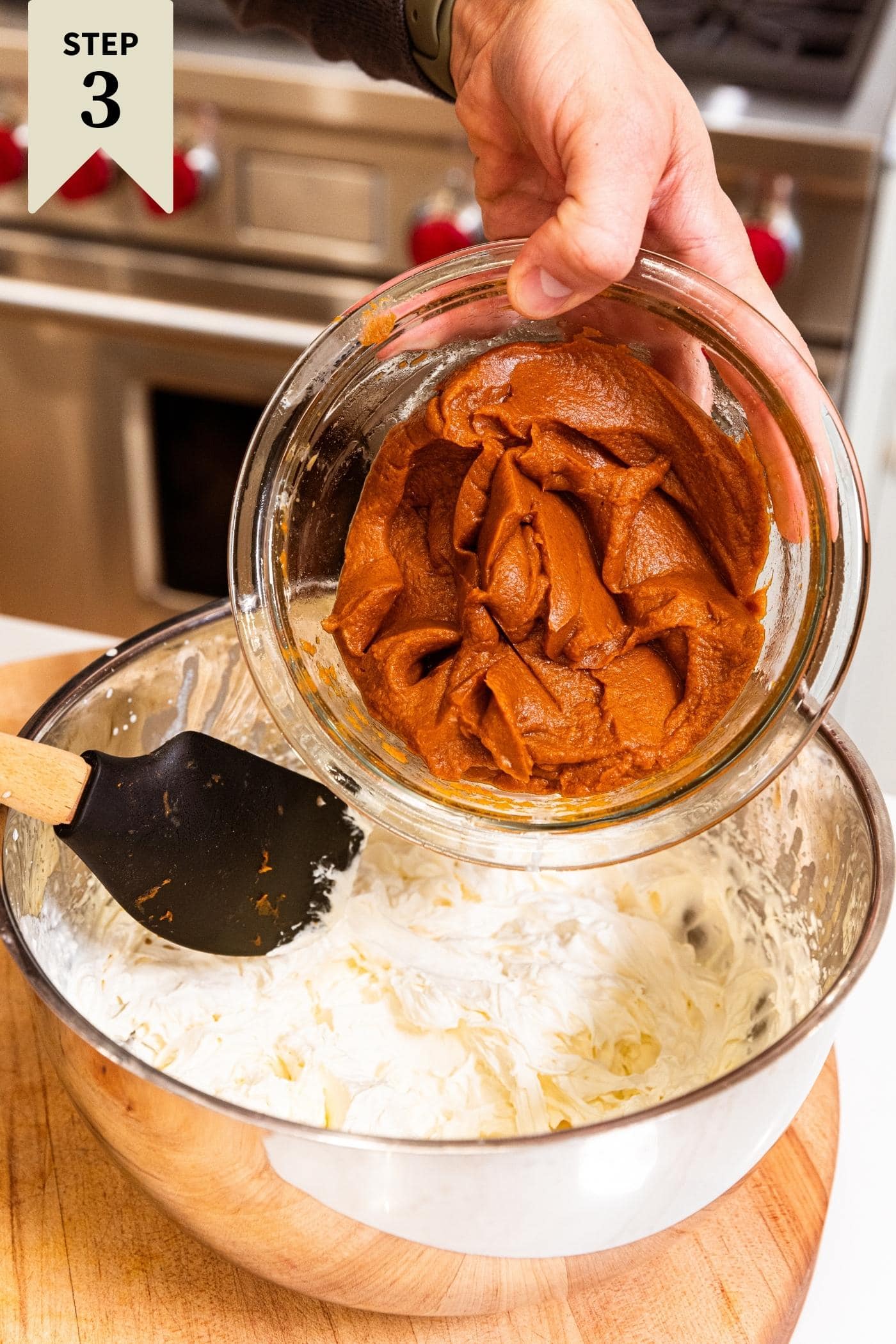 Hand holding glass bowl about to pour pumpkin mixture into the whipped cream in metal bowl with plastic spatula.