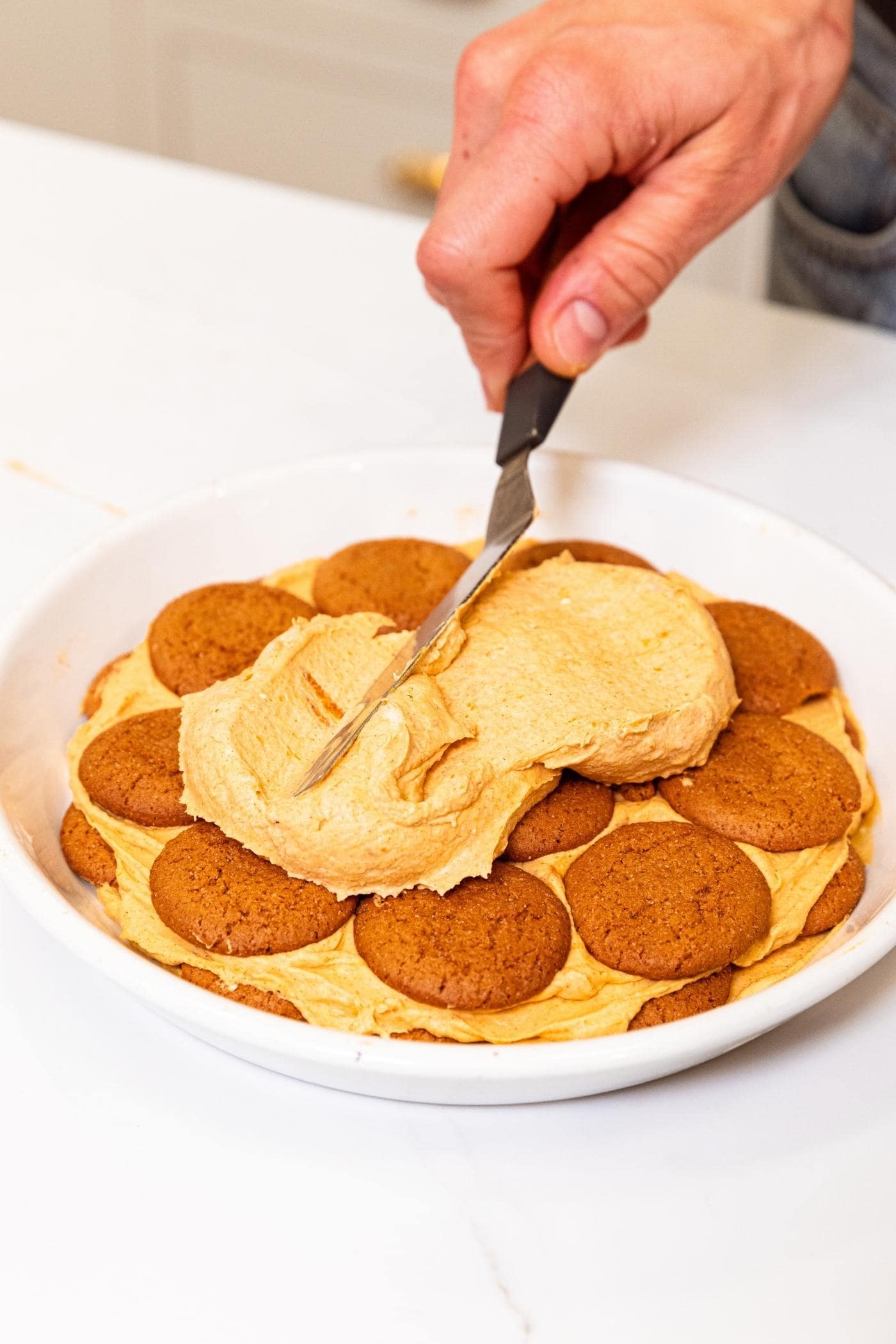 Hand using offset spatula to spread out light brown colored pumpkin whipped cream on top of layer of gingersnap cookies sitting on white plate on white surface.