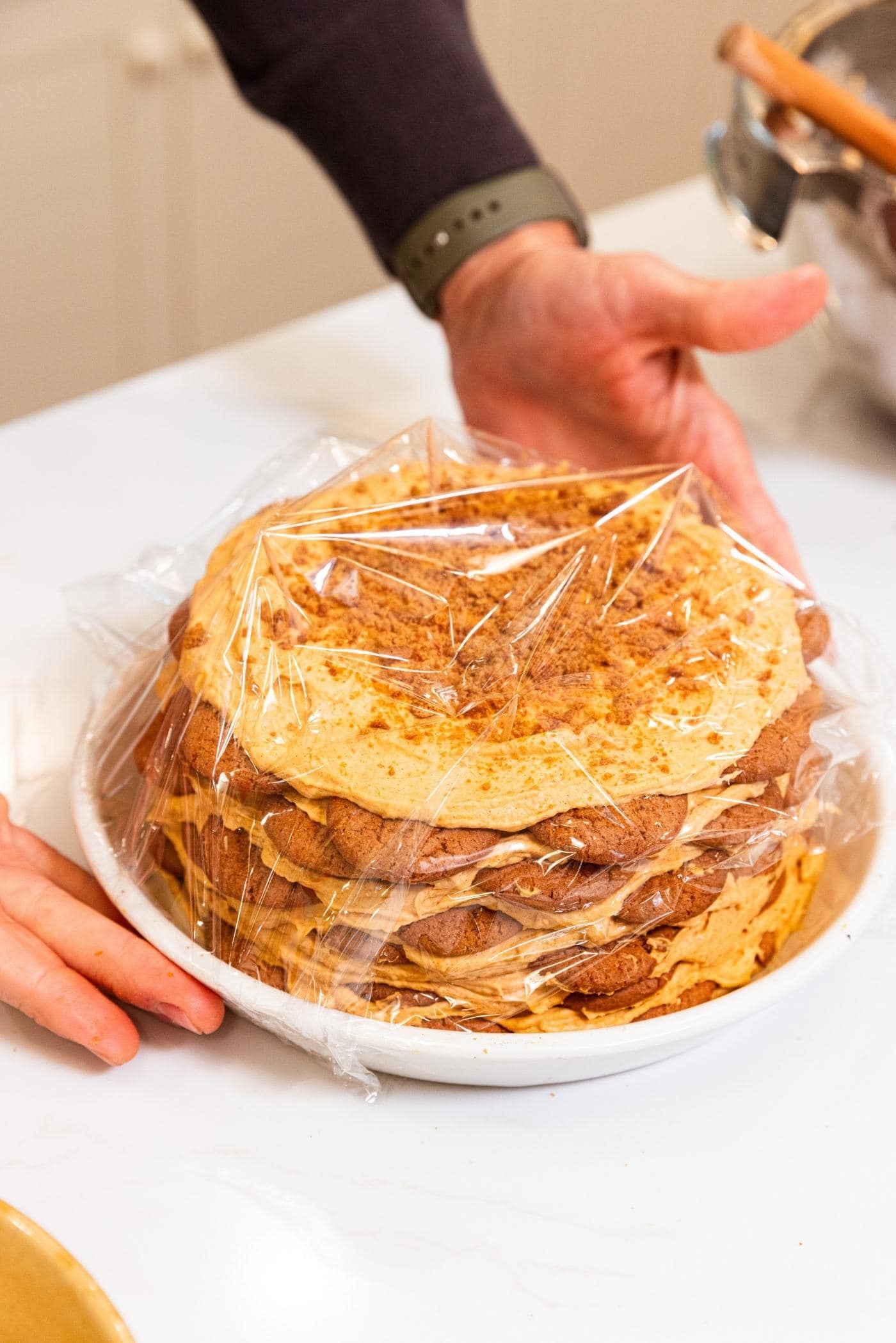 Plastic wrap placed over top of a pumpkin icebox cake with pumpkin whipped cream topping with crumbled gingersnaps on top.