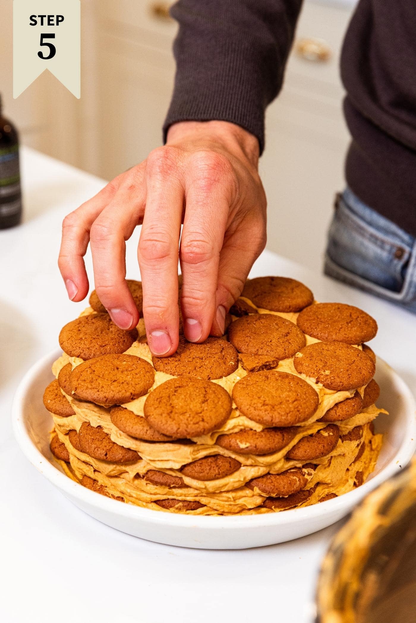 Hand placing last gingersnap cookie on top of cake with white platter underneath.