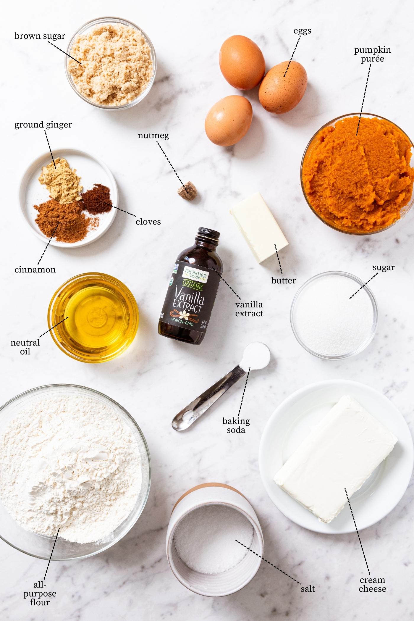 White marble surface filled with all ingredients needed to make a pumpkin coffee cake with cheesecake swirl on top including pumpkin purée, flour, oil, spices, eggs, sugars, vanilla extract, and more.