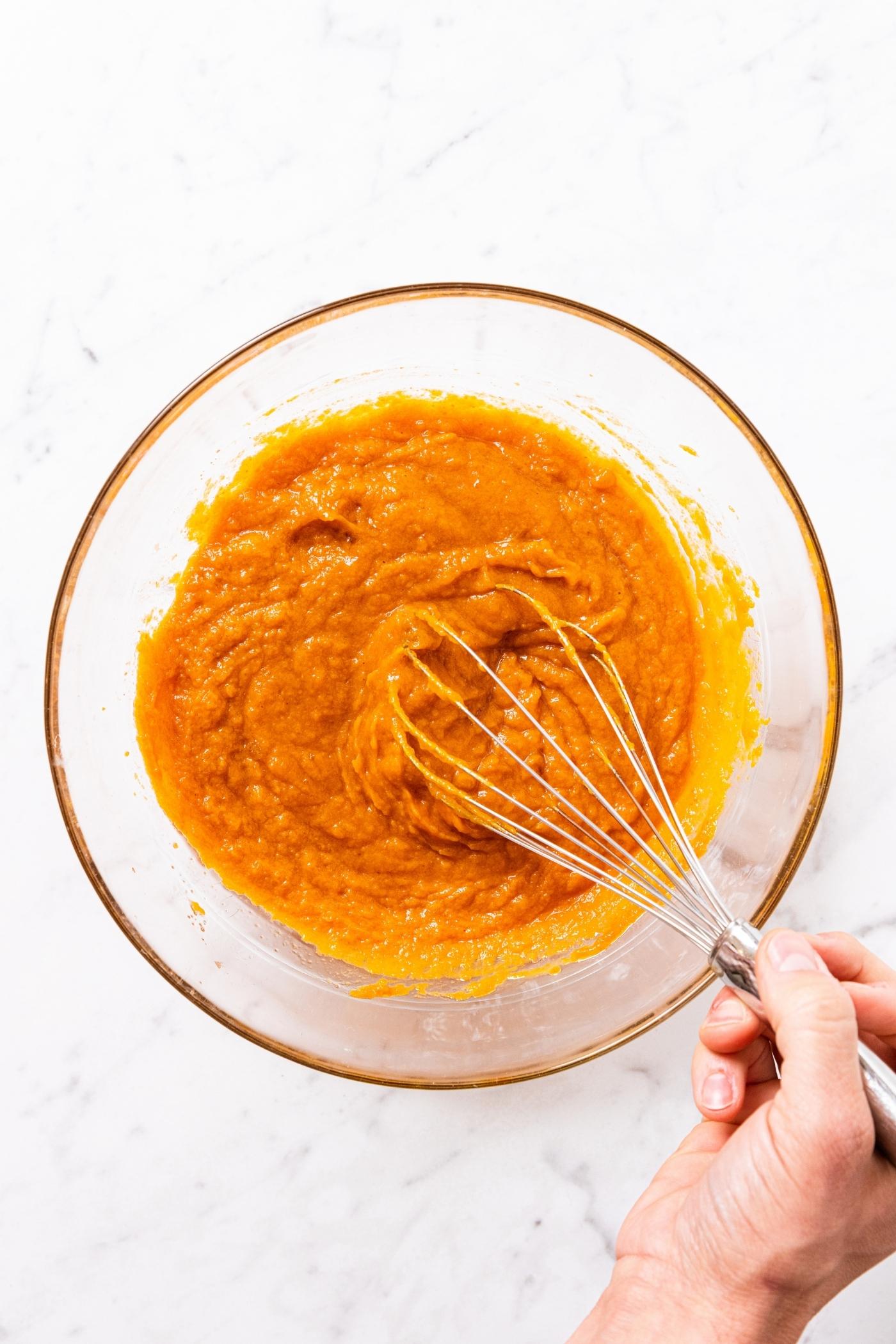 Hand using wire whisk to mix together pumpkin batter in a glass mixing bowl on marble surface.