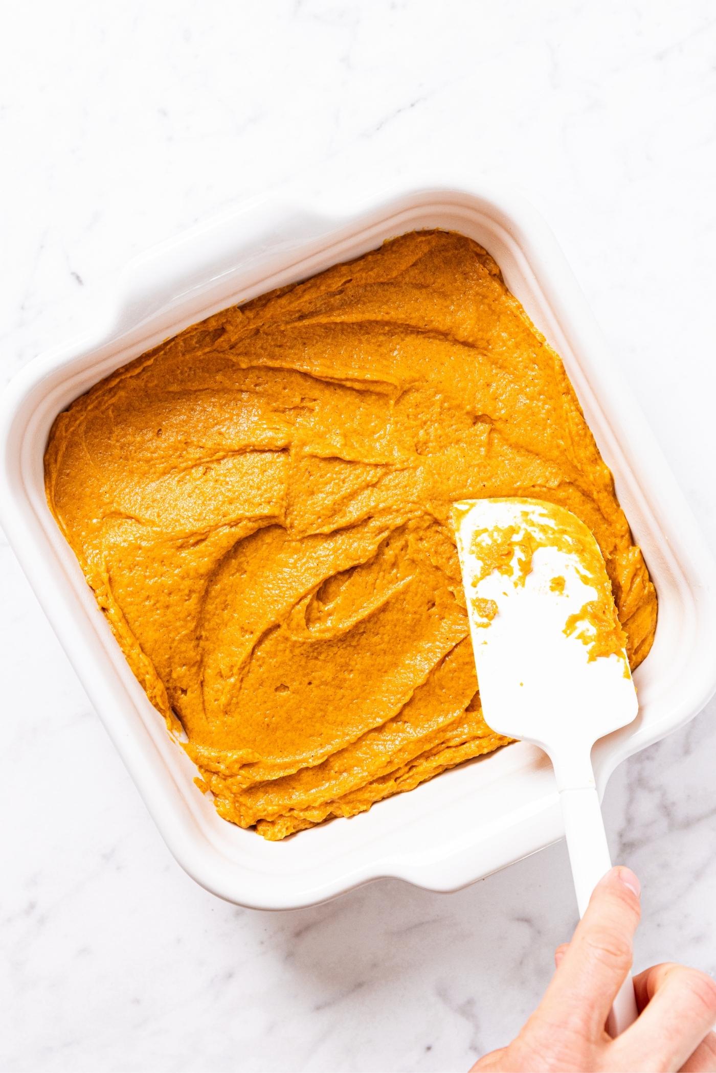 Hand using white spatula to smooth out pumpkin batter in a white baking dish sitting on marble surface.