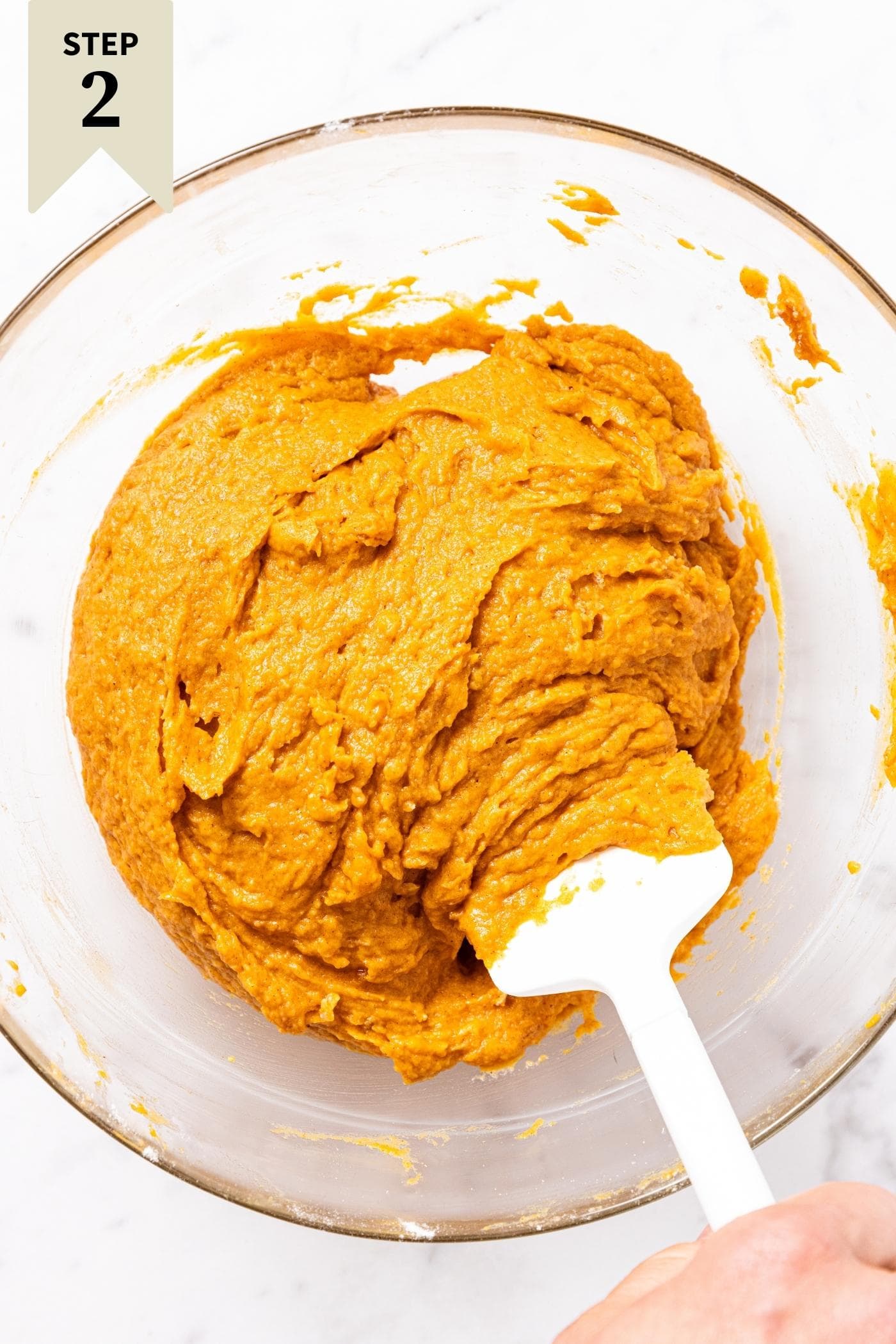Hand using white spatula to mix together pumpkin batter in glass bowl on marble surface.