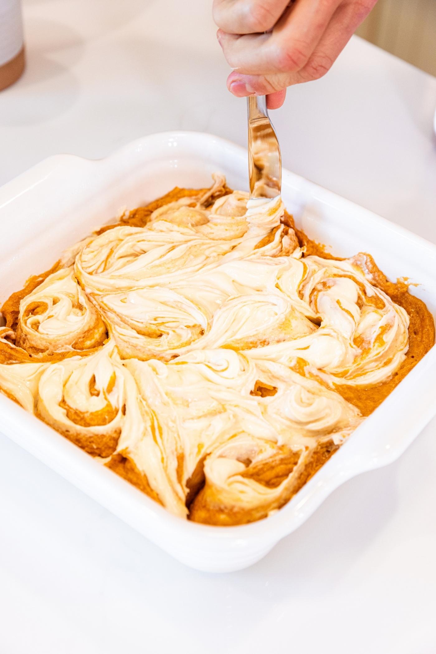 Hand using a table knife to swirl together pumpkin batter with cheesecake mixture in square baking dish on white countertop.