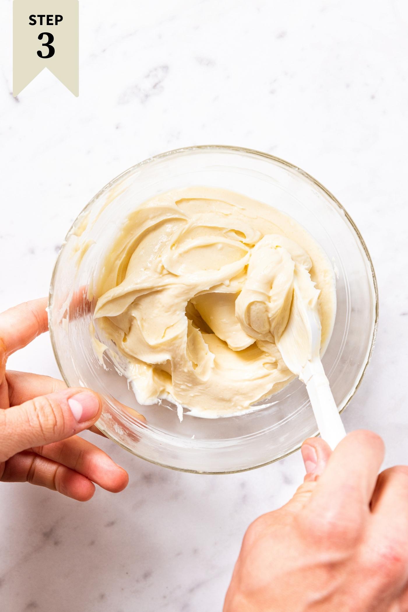 Glass mixing bowl sitting on marble surface with hand mixing together cheesecake swirl mixture with white spatula.