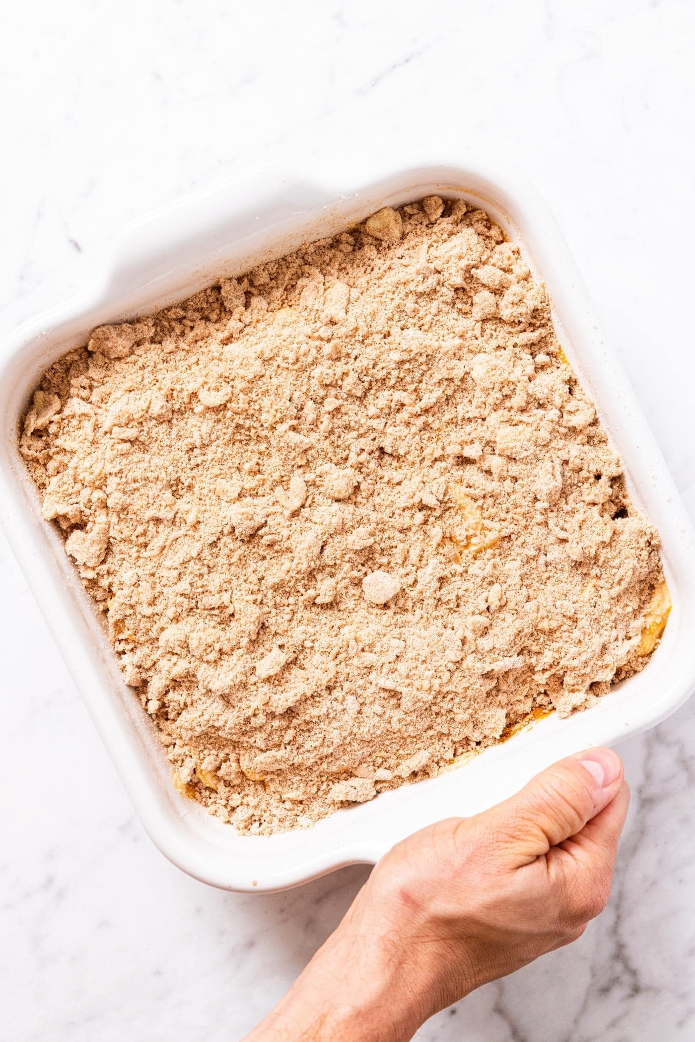 Hand holding white baking dish filled with pumpkin batter topped with streusel topping before going into oven to bake.
