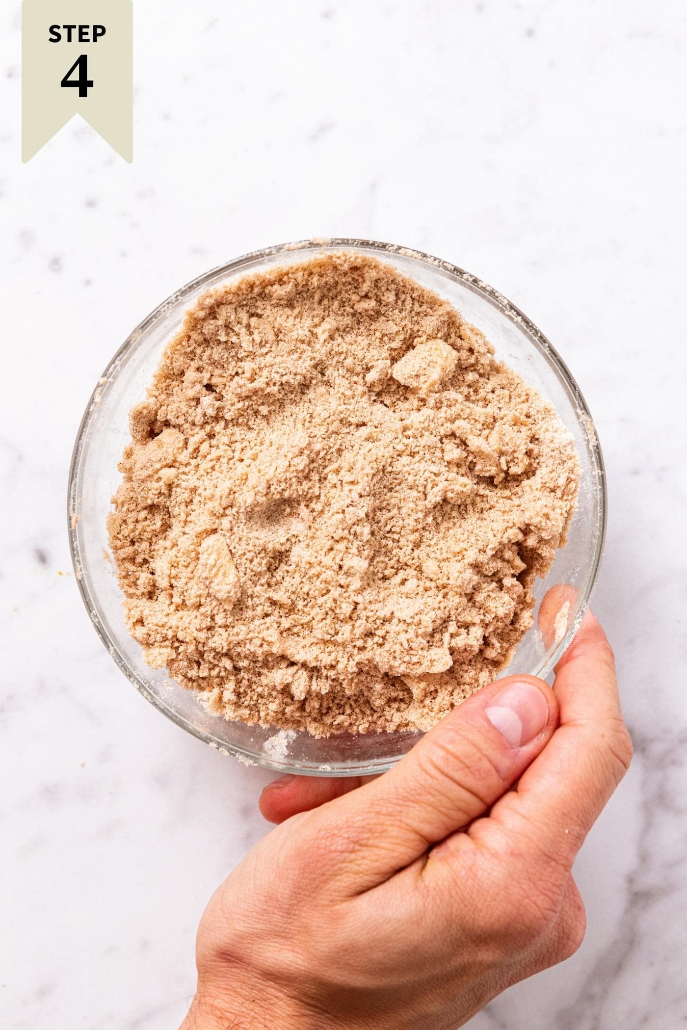 Hand holding side of small glass bowl filled with crushed up sand-like streusel topping before being sprinkled on top of a cake.