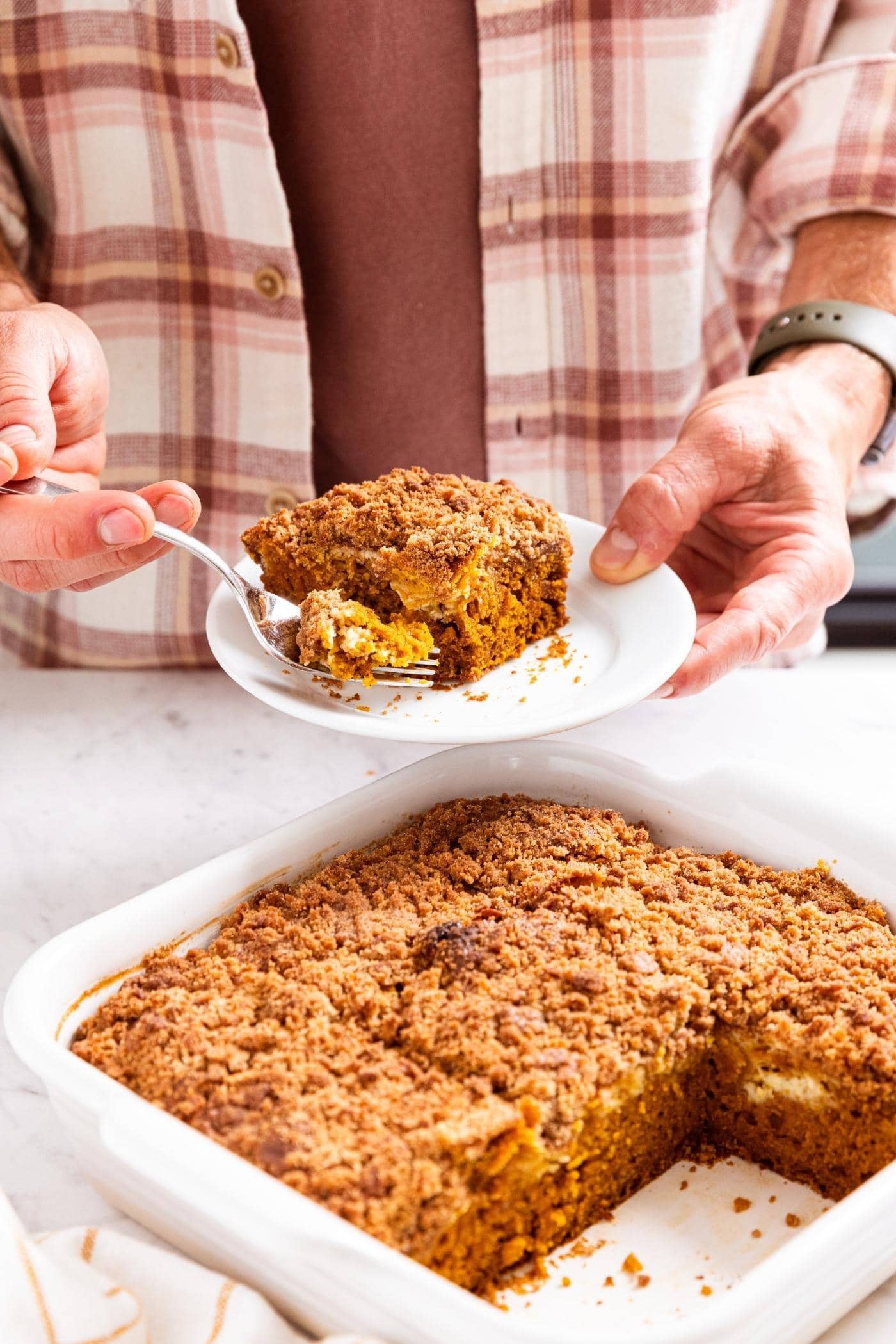 Hand holding small white plate with square slice of pumpkin coffee cake with rest of cake in white baking dish below.