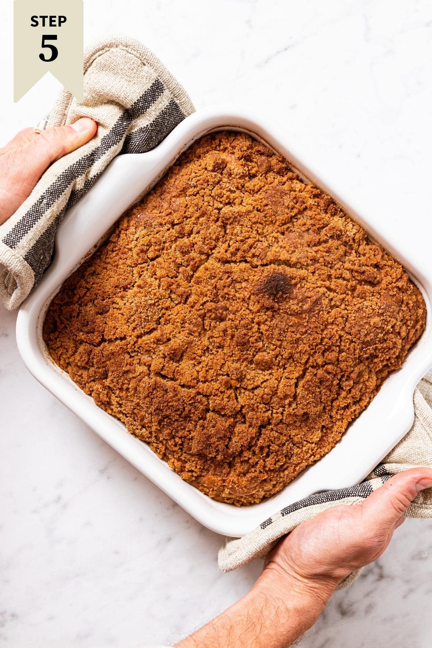 Hand holding white baking dish with potholders filled with baked pumpkin coffee cake with streusel topping.
