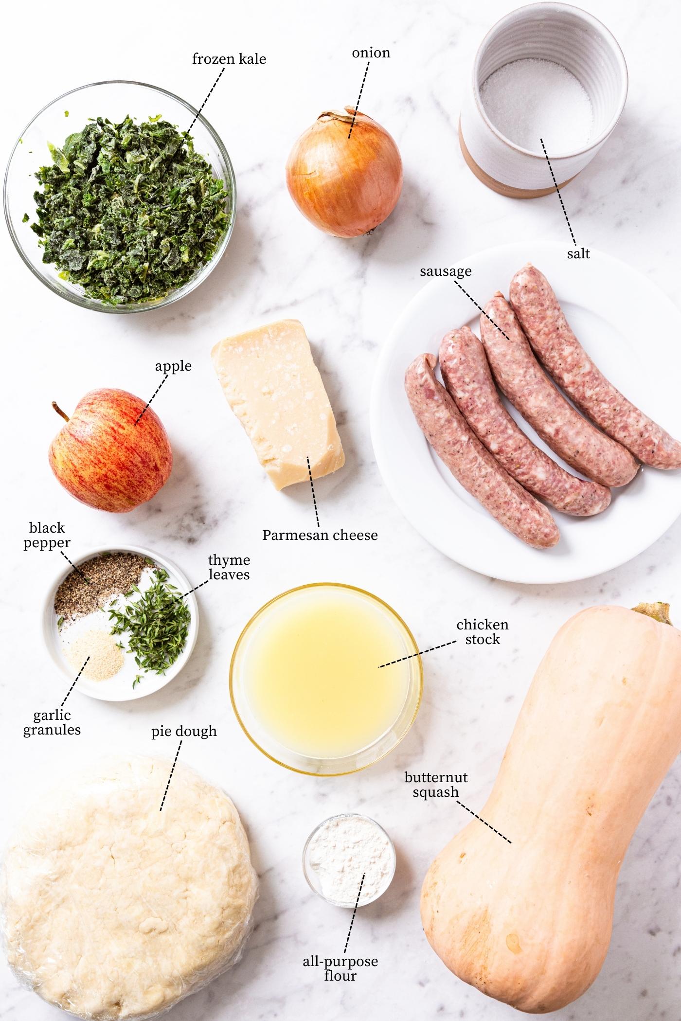 Marble surface filled with all of the ingredients needed to make savory squash hand pies including butternut squash, sausage, kale, pie dough, spices, and more.