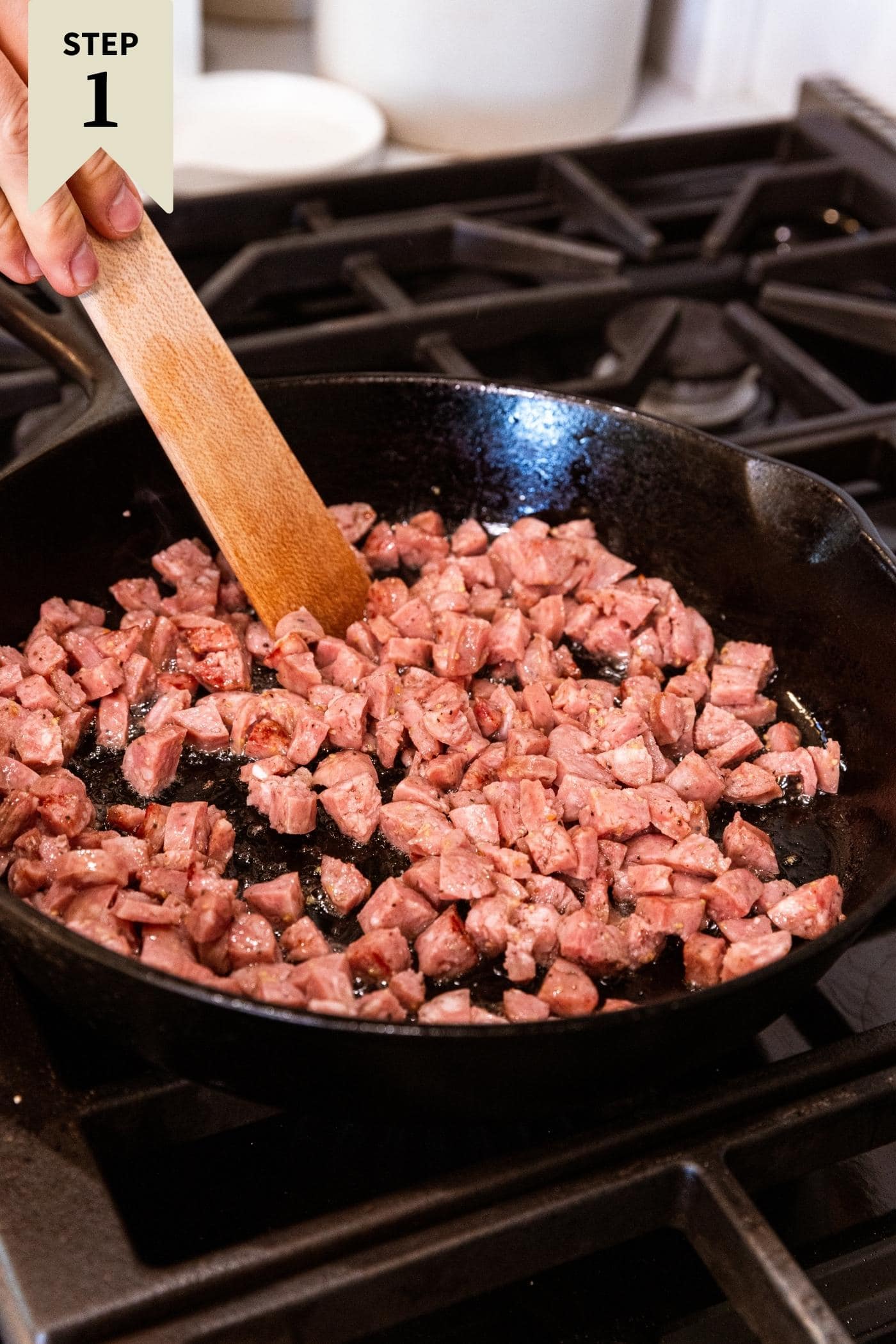 Wooden spatula stirring sausage cut into small diced squares in black cast iron skillet on stovetop.